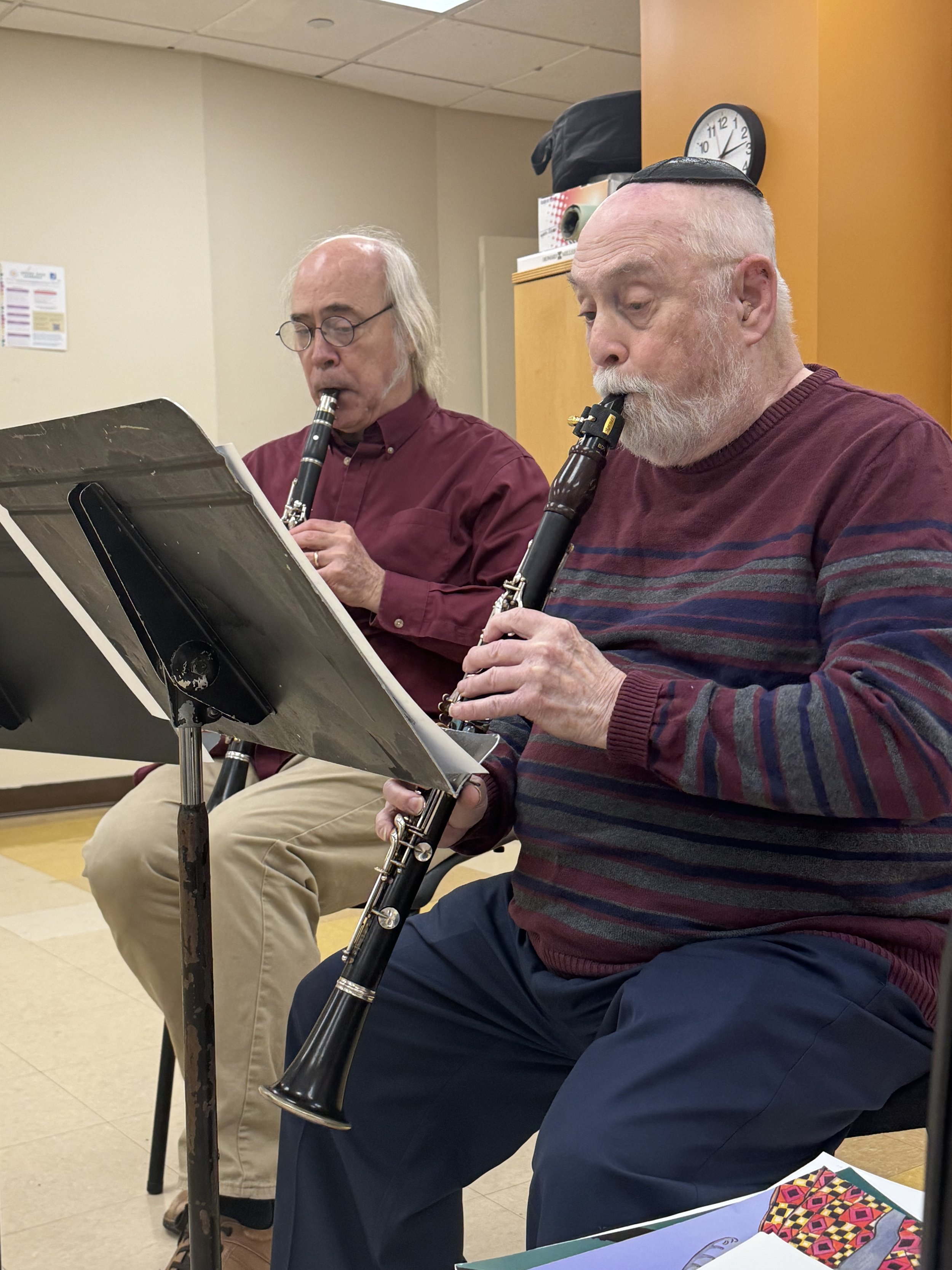 Two elderly men playing recorders while sitting at music stands in a room. One man has long gray hair and glasses, the other has a beard and is wearing a yarmulke and a striped sweater.