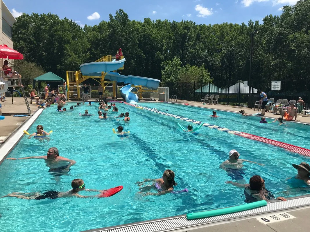 People swimming and playing in an outdoor swimming pool with a water slide, surrounded by trees and poolside chairs.
