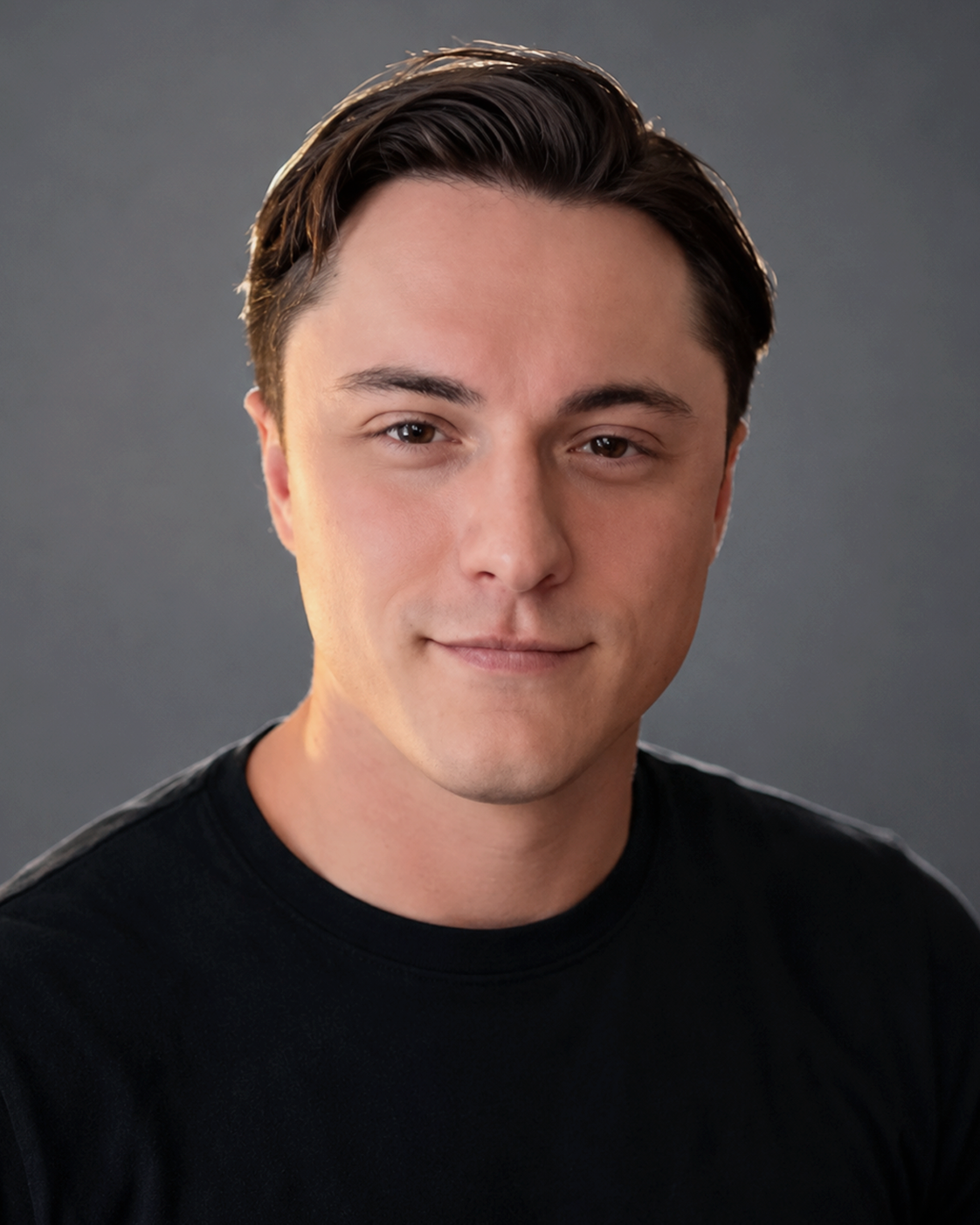Portrait of a young man with dark hair and fair skin, wearing a black shirt, smiling slightly, against a gray background.