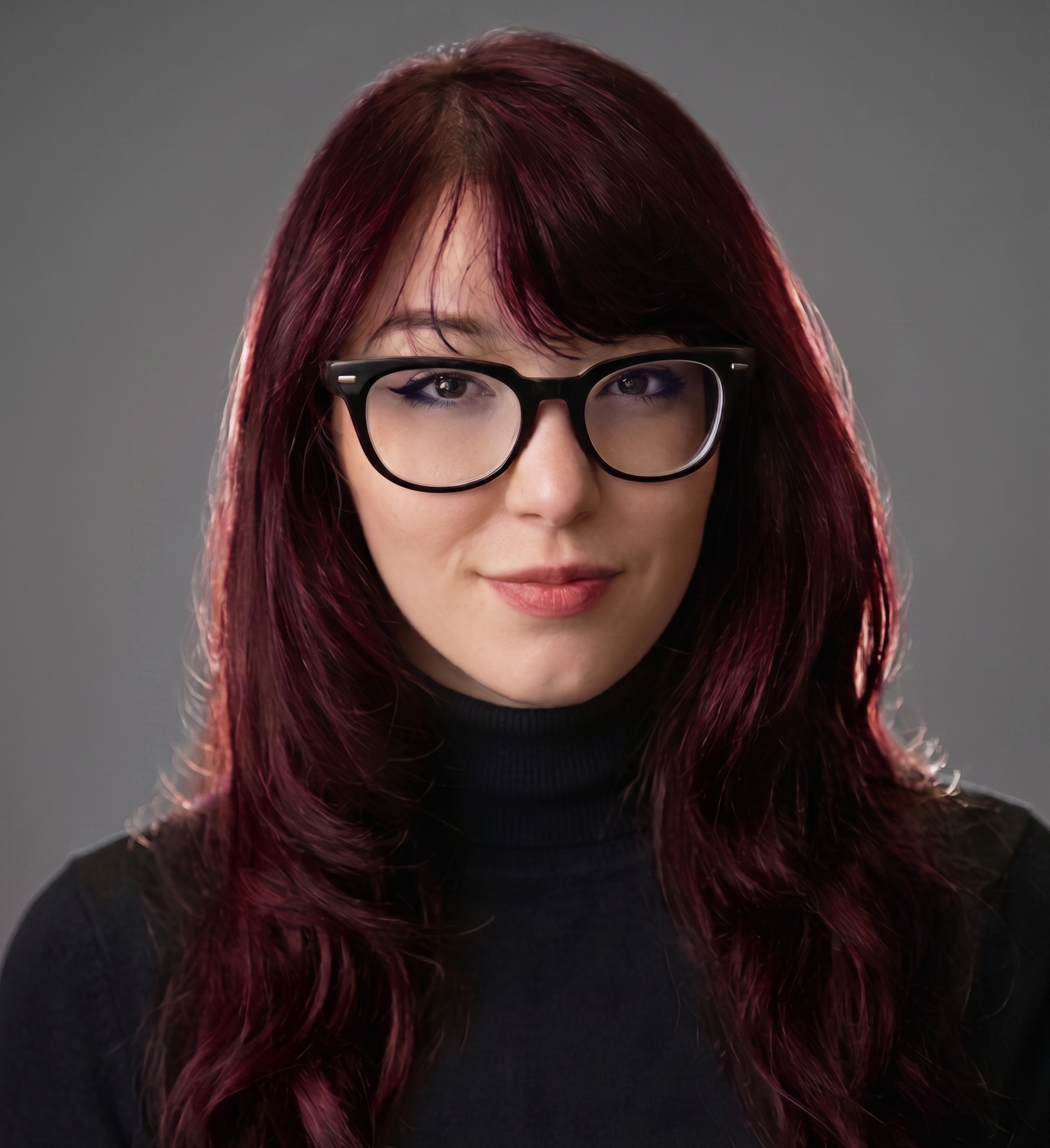 Close-up of a woman with long, dark red hair, wearing glasses, and dressed in a black top, against a neutral gray background.