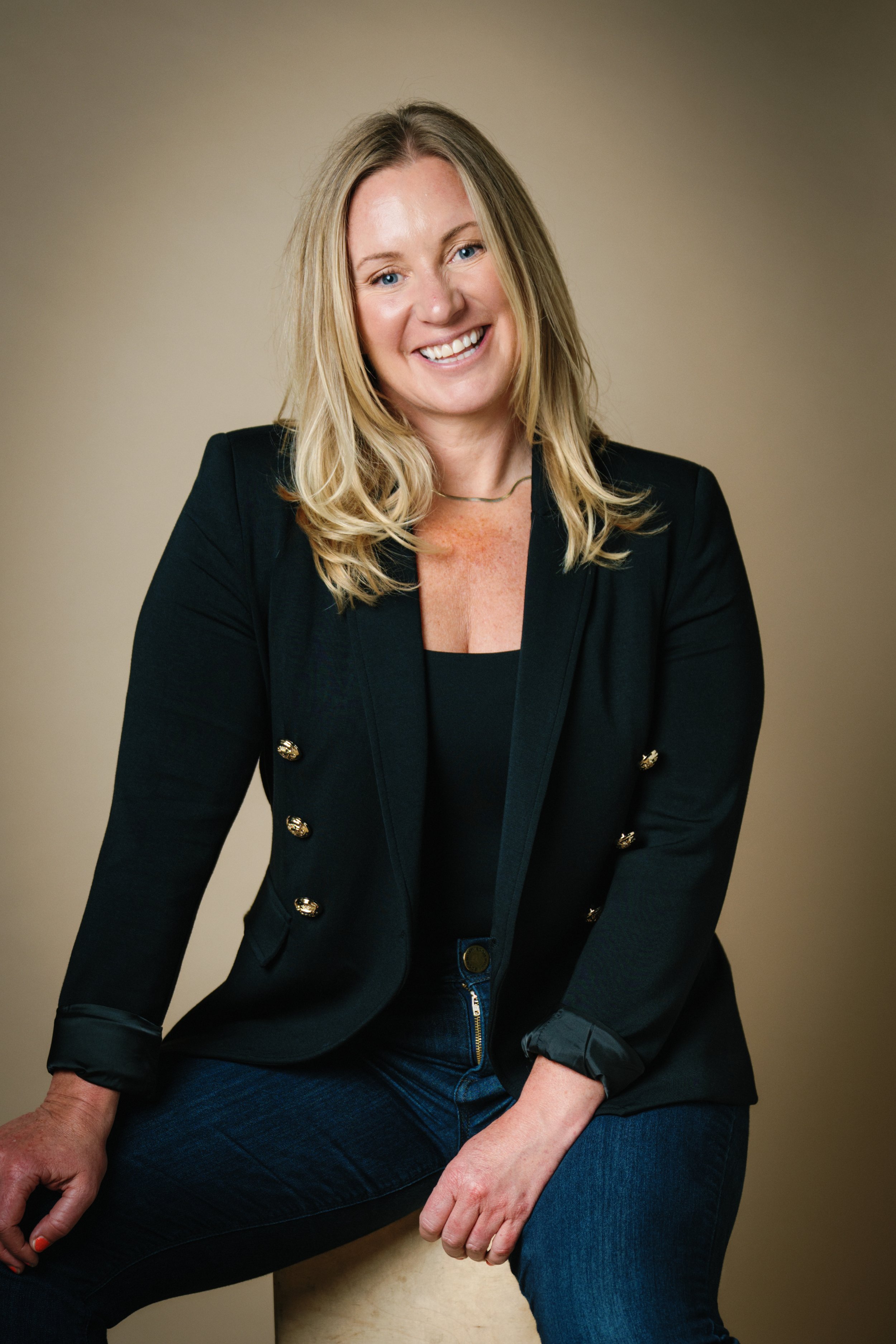 A woman with blonde hair wearing a black blazer and blue jeans, sitting on a beige stool against a beige background, smiling at the camera.