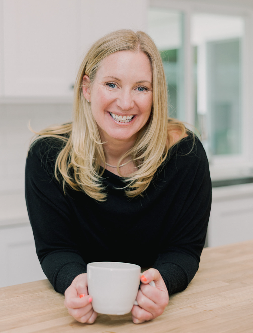 A smiling woman with blonde hair and blue eyes holding a white coffee mug in a kitchen setting.