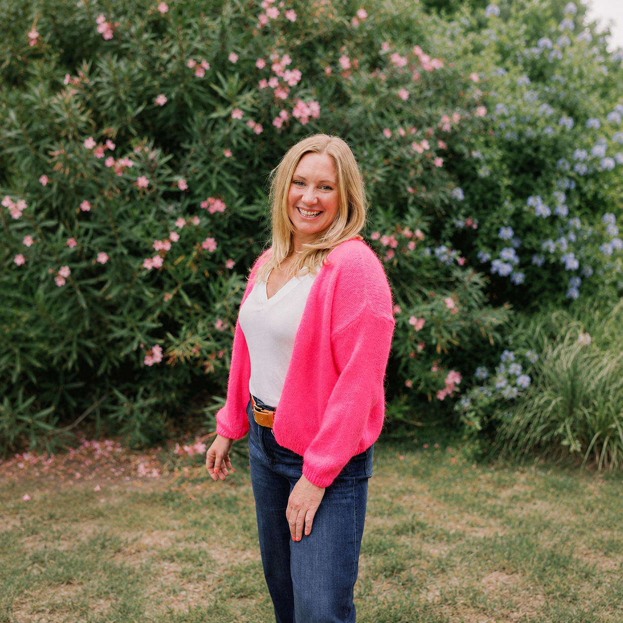 A woman smiling outdoors in front of flowering bushes, wearing a pink cardigan, white top, and jeans.