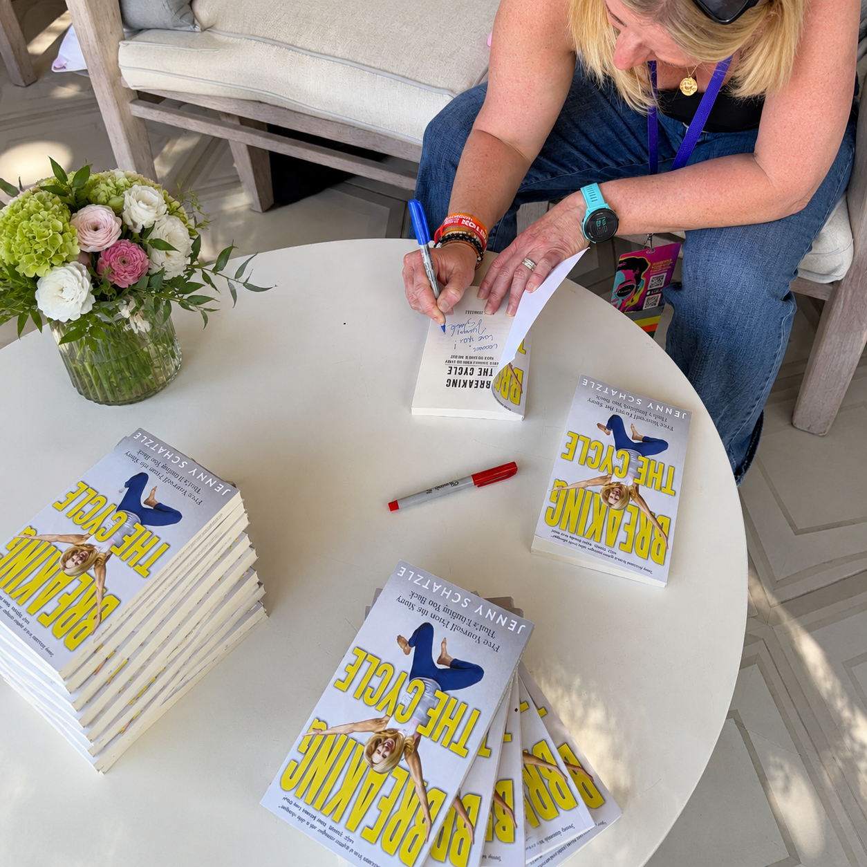 A woman sitting at a white round table signing a book titled 'Breaking the Cycle' by Jenny Schartz. The table has multiple copies of the book, a red marker, and a vase of pink and white flowers. The woman is wearing a black top, jeans, a watch, bracelets, and black sunglasses on her head.