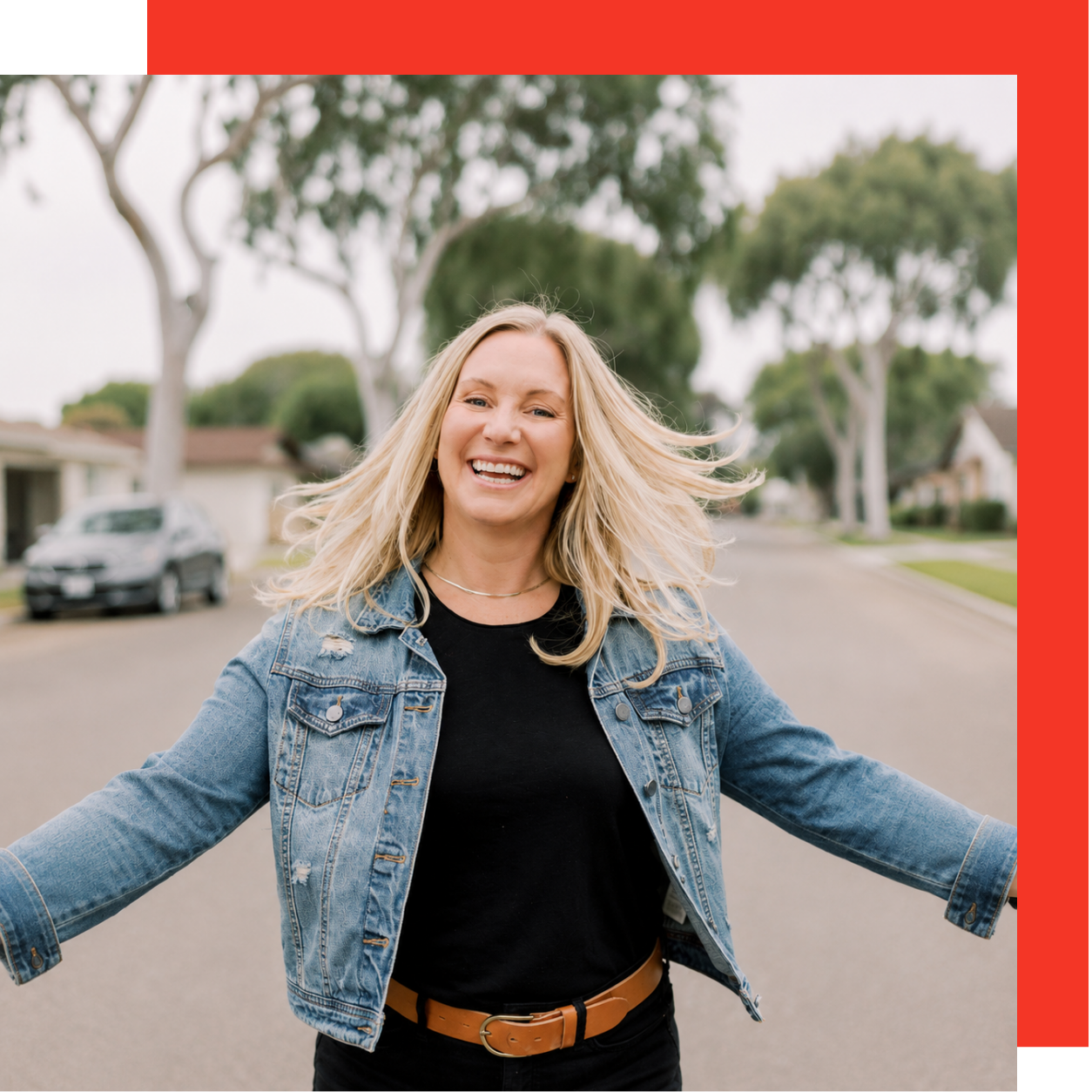 A cheerful woman with blonde hair, wearing a denim jacket and black top, standing in the middle of a suburban street with trees and houses in the background.