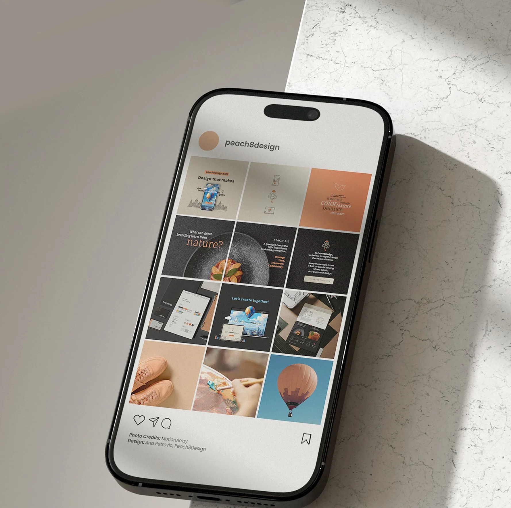Smartphone displaying an Instagram profile page for peacht8design with multiple posts including a phone with a waterscape, a rocket, a bowl of food, and a hot air balloon, resting on a white surface.