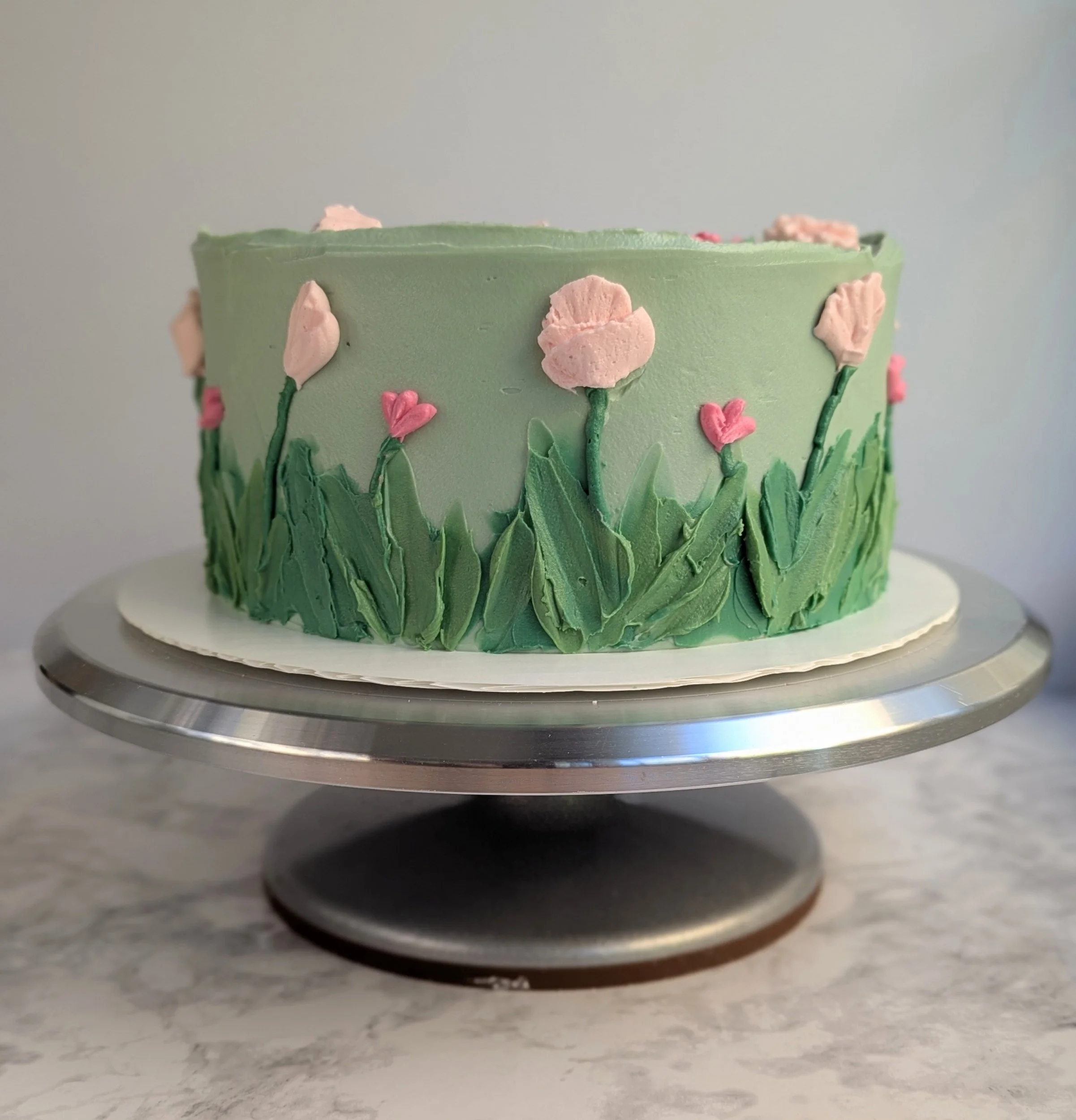 A round, layered cake with green and pink floral decorations on a silver cake stand.