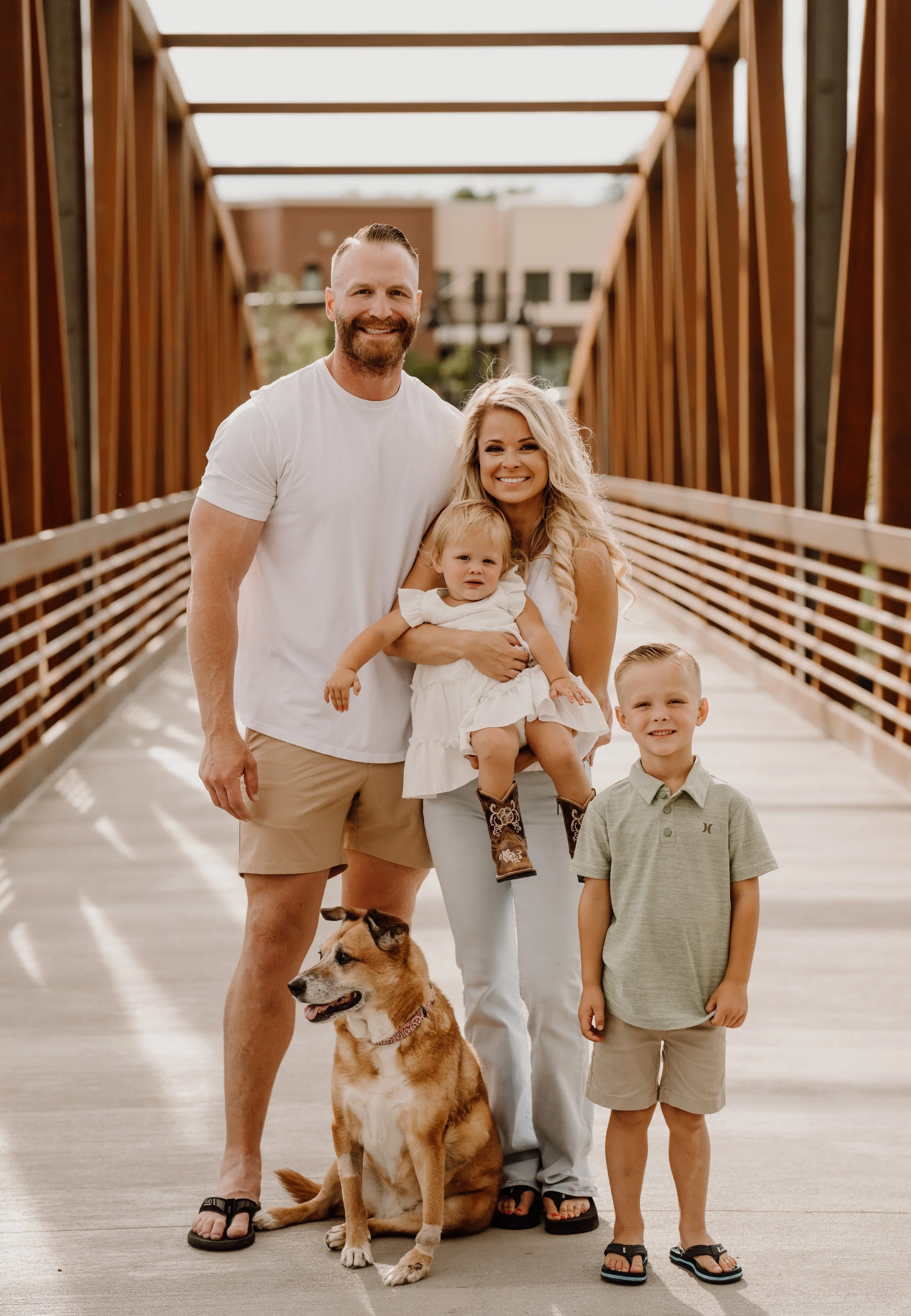 A family portrait on a bridge with two adults, two children, a dog, and a puppy, all smiling.