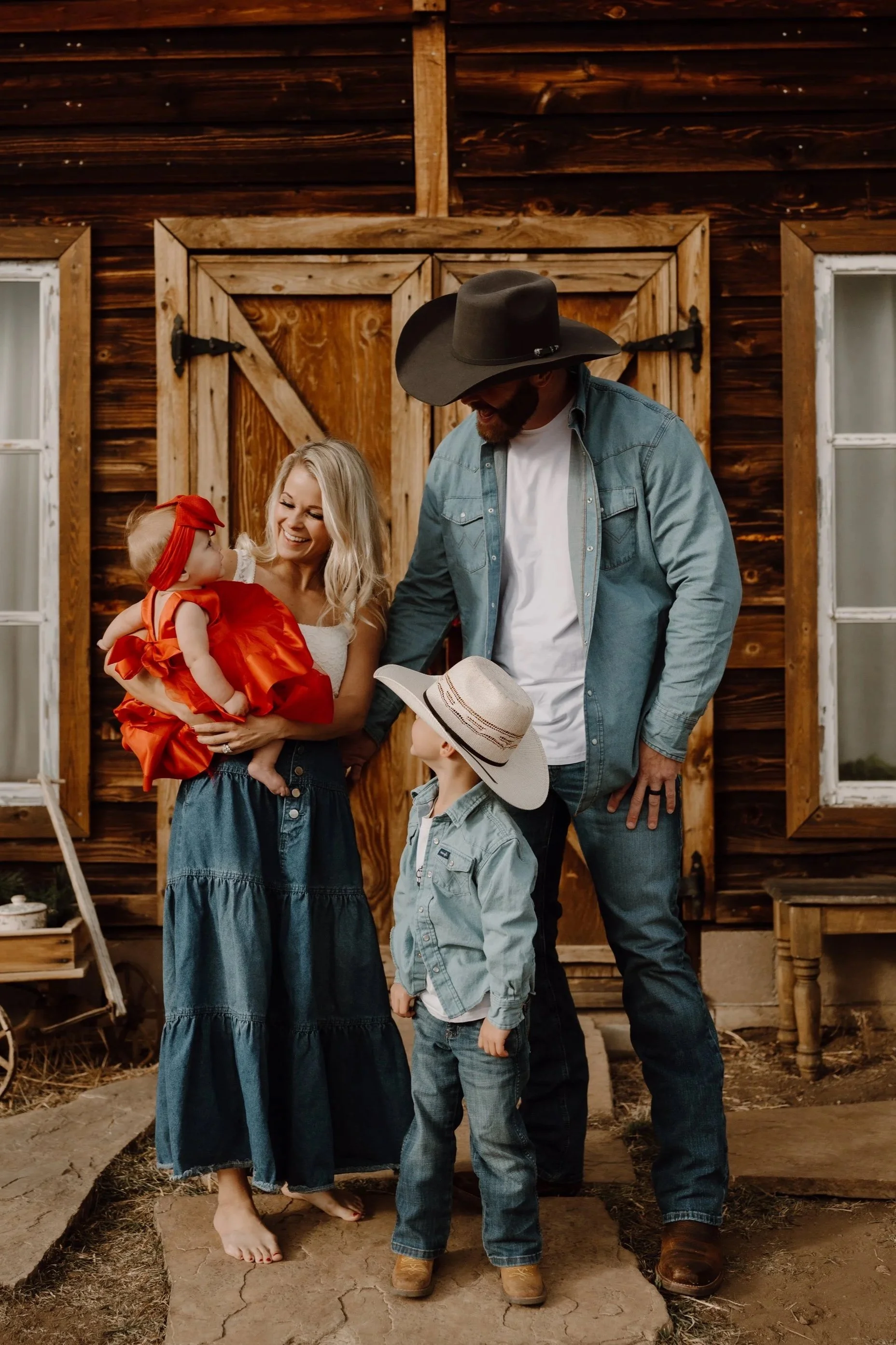 A family of four standing outside in front of a wooden barn. The woman is holding a baby girl dressed in red, and there are two young boys standing nearby. The man is wearing a large cowboy hat and a denim jacket, looking at a book. The scene has a rustic, country vibe.