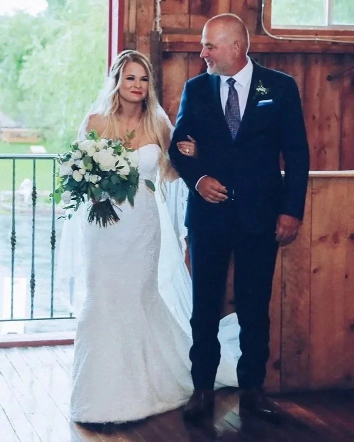 A bride in a white wedding dress holding a bouquet of white flowers walking down the aisle with her father, who is dressed in a dark suit, inside a wooden barn.