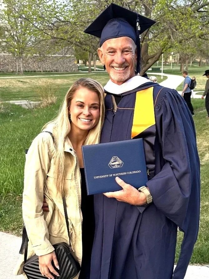 A graduation ceremony with a man in cap and gown holding a diploma from the University of Northern Colorado, standing outdoors on a university campus, smiling with a young woman in casual attire.