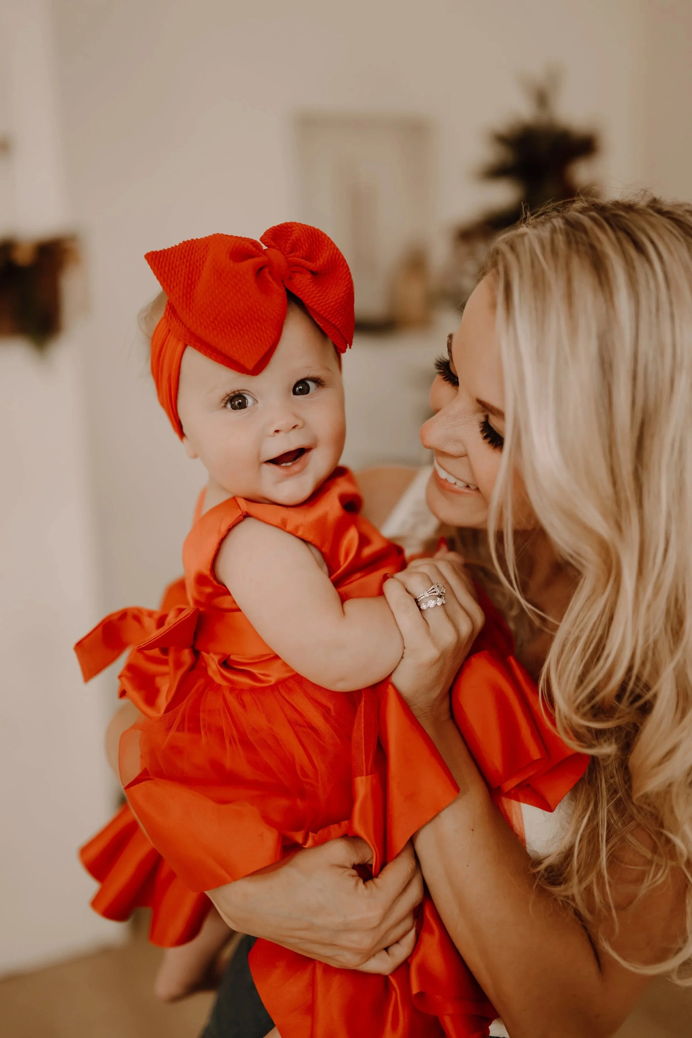 A woman with long blonde hair and a bright, wide smile holds a baby girl dressed in an orange dress with a large matching orange bow on her head. The baby is looking at the camera with a happy expression.