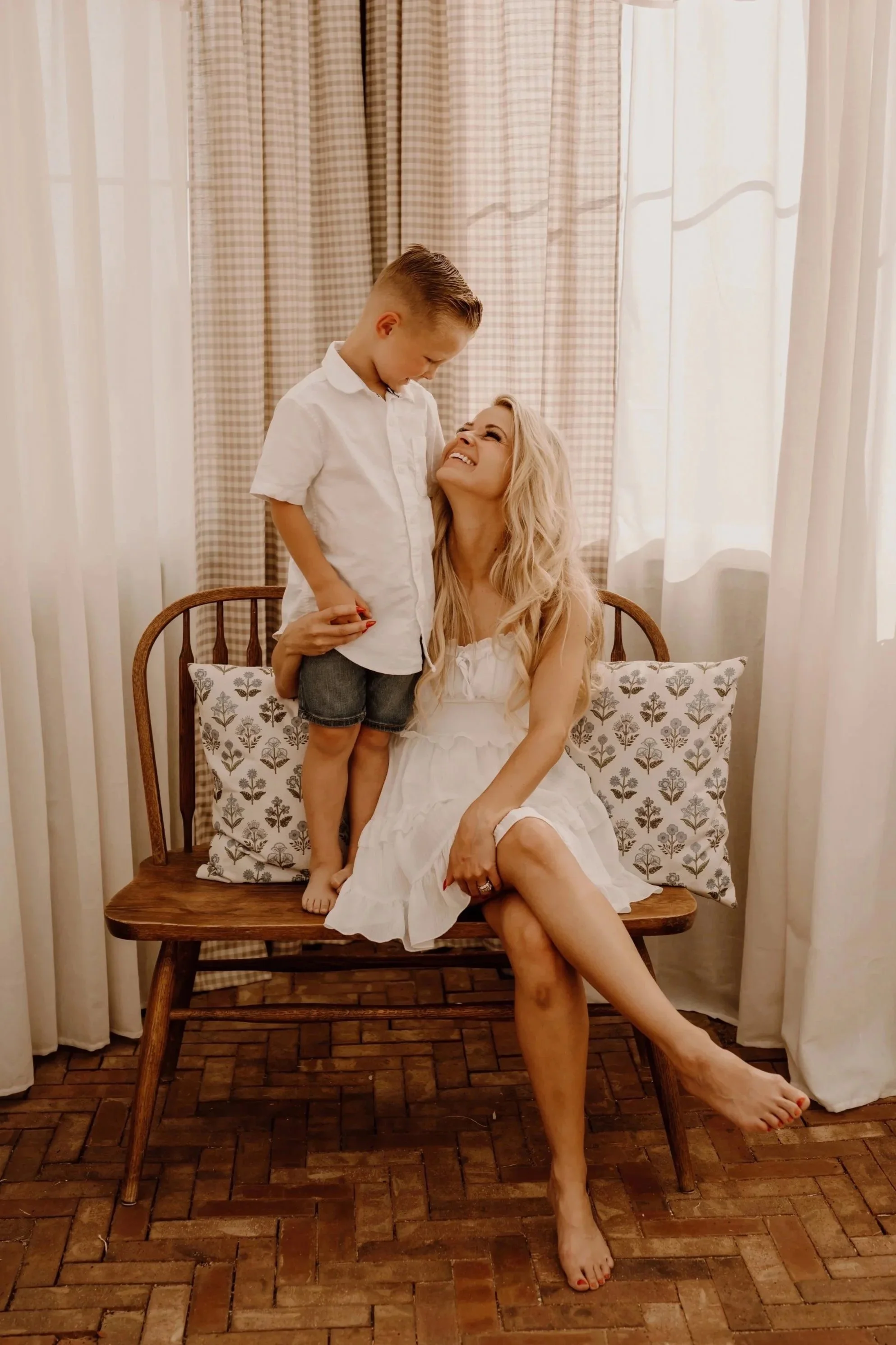 A woman sitting on a wooden bench with a young boy standing on her lap, both smiling and looking at each other, in a room with soft lighting and cream-colored curtains.