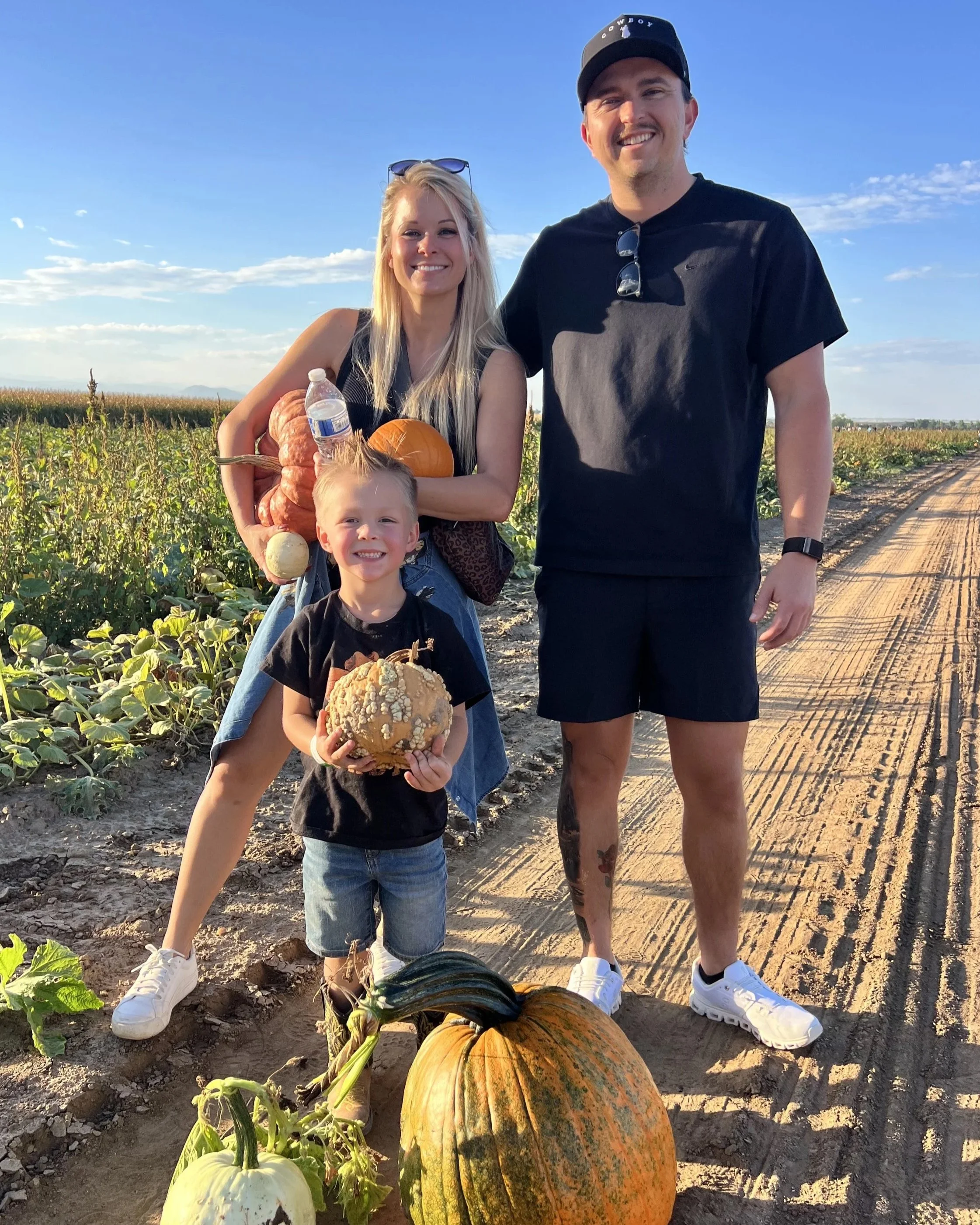 A family of three standing in a pumpkin patch during the day. The woman is holding a bottle of water and a pumpkin, and the young boy is holding a large decorative pumpkin. The man stands next to them, all smiling at the camera. The sky is clear with some clouds, and the pumpkin patch stretches into the distance.