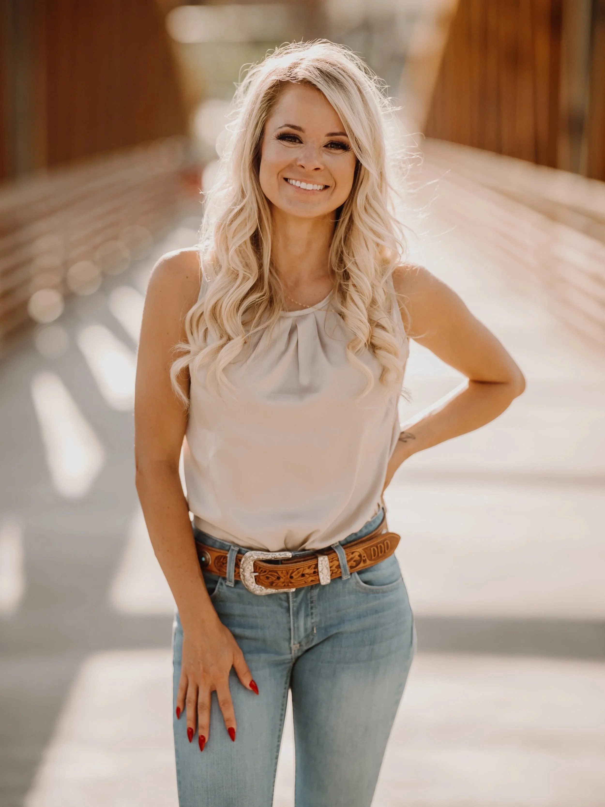 A smiling blonde woman wearing a white sleeveless top and light blue jeans, standing outdoors in front of a wooden bridge, with her right hand on her hip and red painted nails.