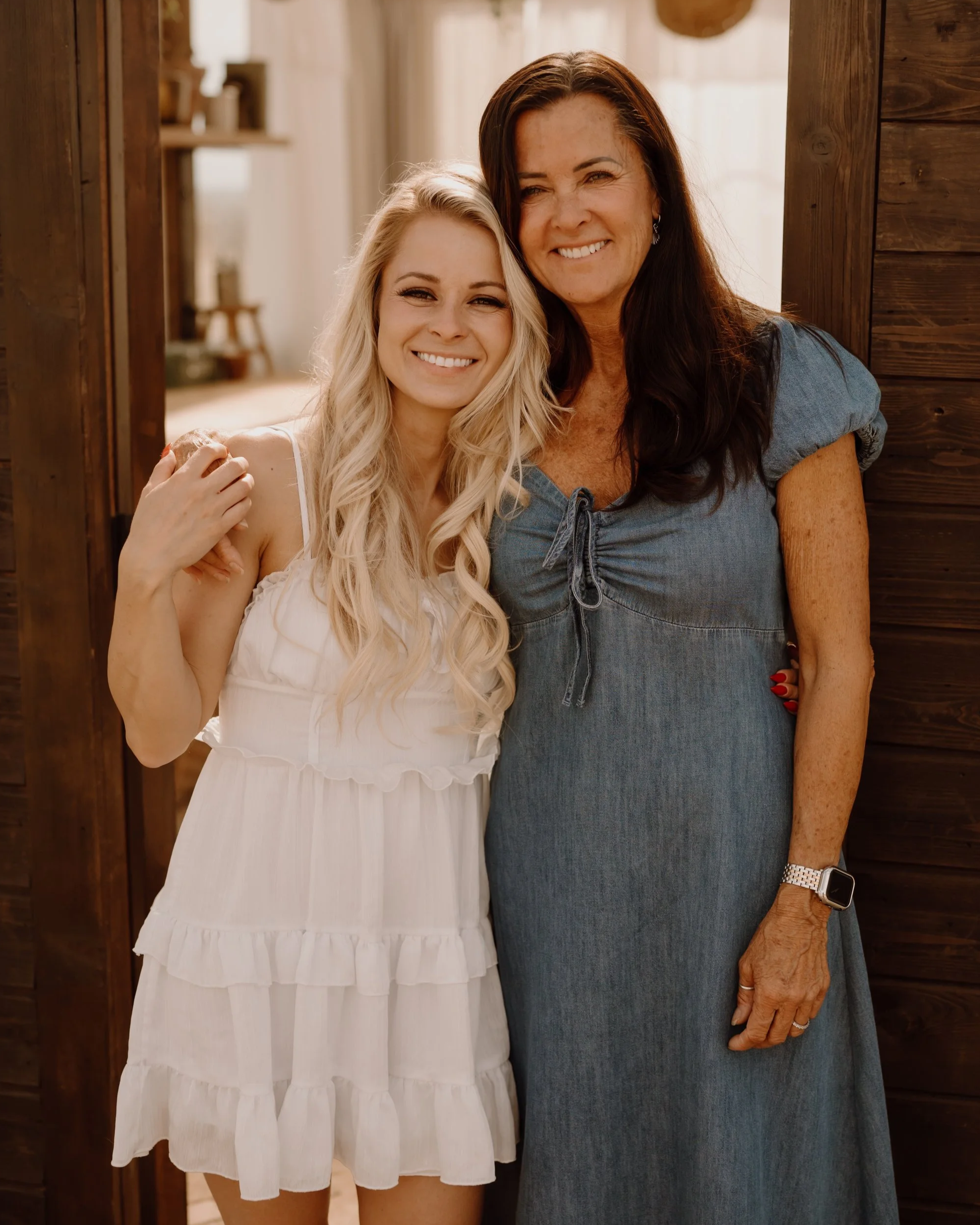 Two women smiling and posing together in front of a wooden doorway. The woman on the left has long blonde hair and is wearing a white dress, while the woman on the right has long dark hair, is wearing a blue dress, and has an Apple Watch on her wrist.