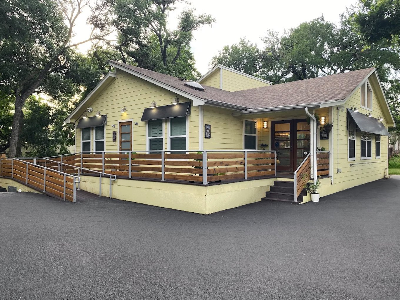Yellow commercial building with multiple windows, black awnings, and outdoor lighting, featuring a wooden ramp and small stairs at the entrance, surrounded by trees.