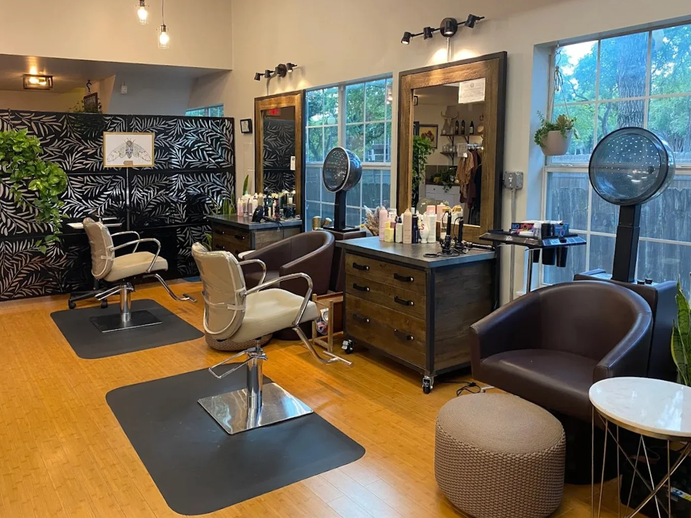 Interior of a hair salon with styling chairs, mirrors, hair drying stations, and decorative plants, illuminated by natural light through large windows.