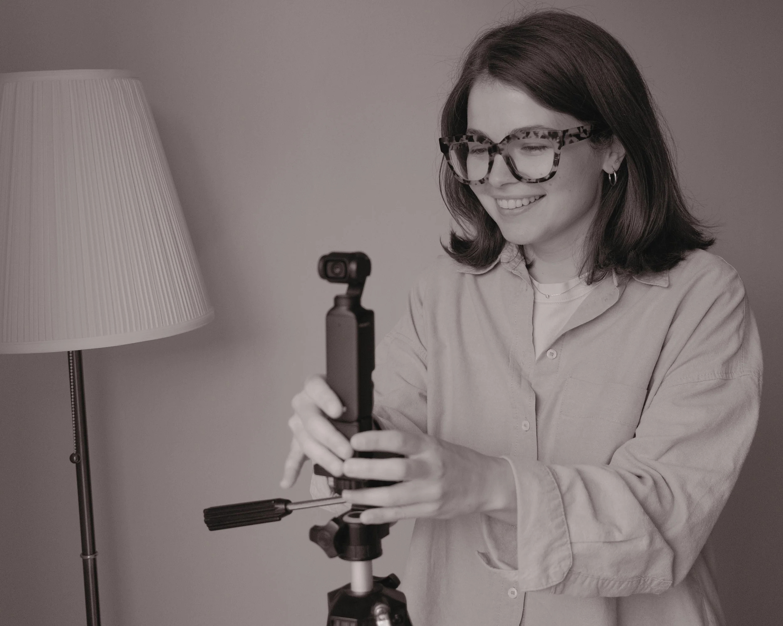 A woman with dark hair, wearing glasses and earrings, smiling and holding a camera, standing next to a lamp.