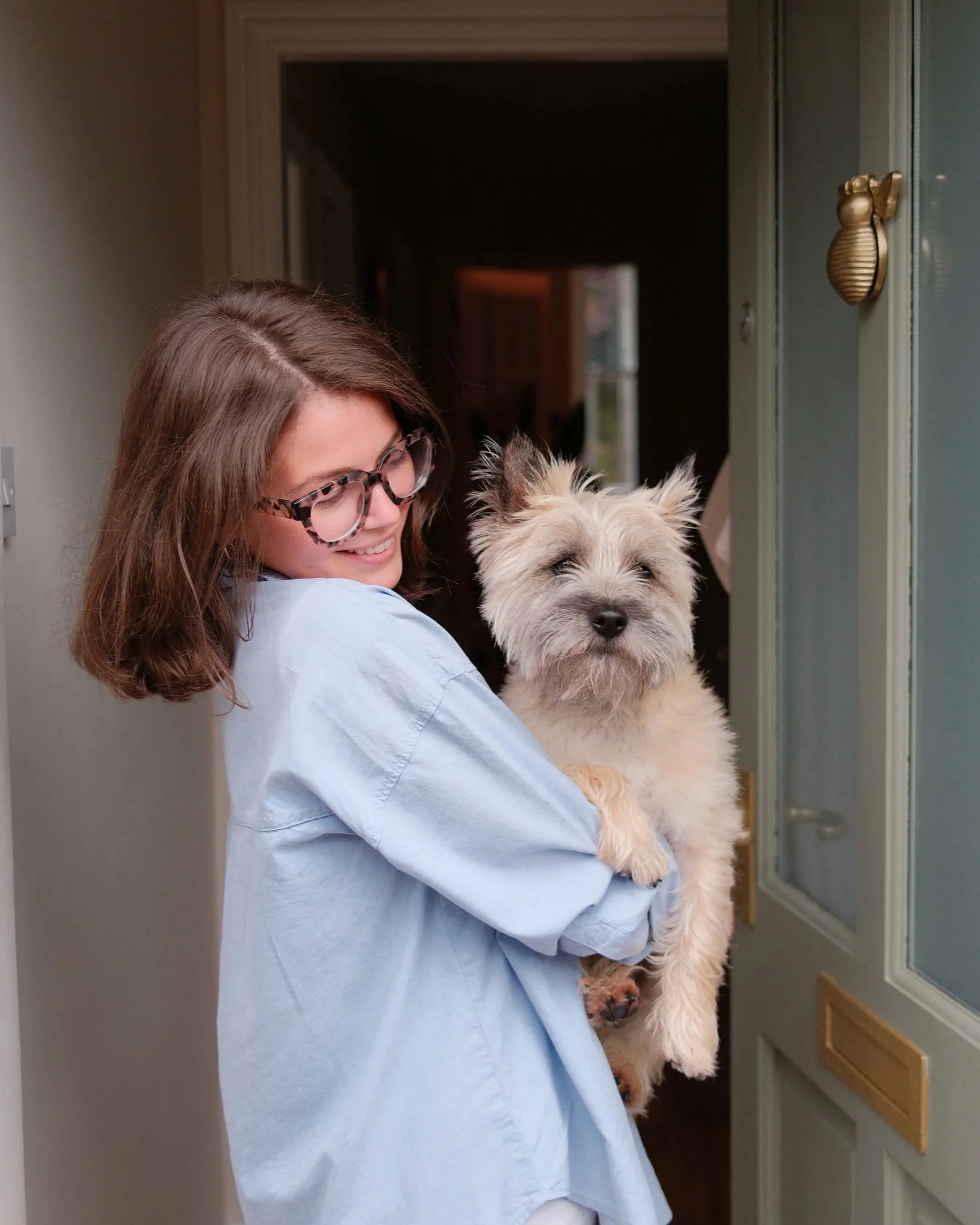 A woman with brown hair and glasses smiling while holding a fluffy dog in front of a house door.