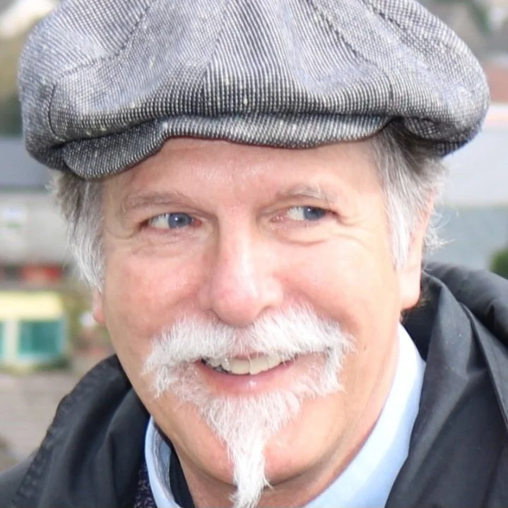Close-up of Tom Laramine with a gray beard and mustache, wearing a gray flat cap and a black jacket, smiling outdoors.