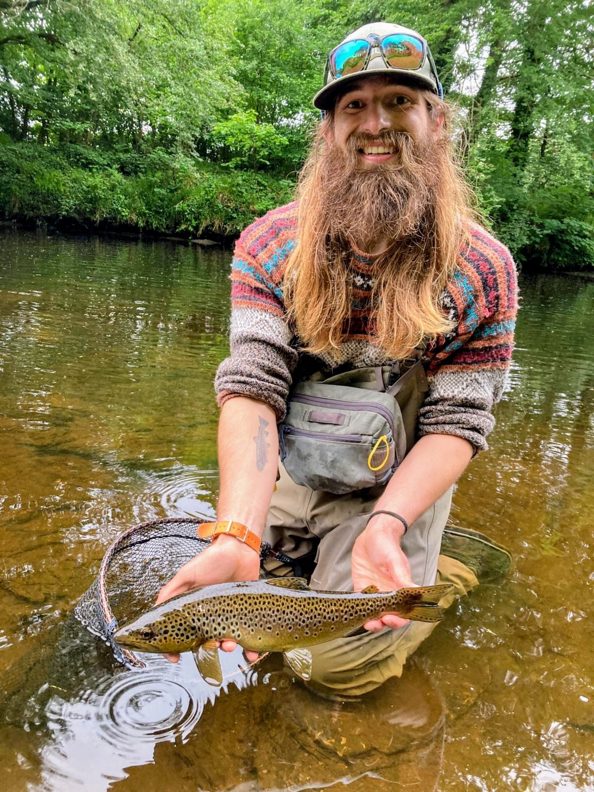 A man with a beard, wearing a cap and sunglasses, stands in a shallow river, holding a brown spotted fish with both hands, surrounded by green trees.