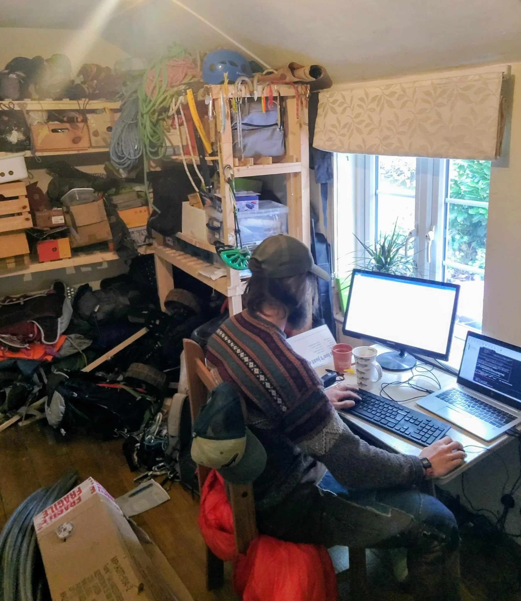 A person using a computer station in a cluttered room with boxes, cables, and tools, with natural light coming through a window covered by a curtain.
