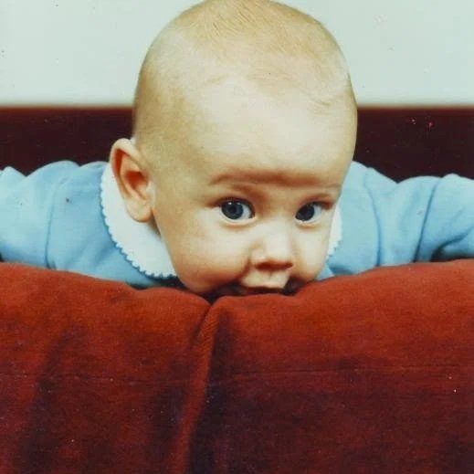 A baby with a bald head and big blue eyes leaning over the edge of a red couch, wearing a light blue outfit with a white collar. Brice Williams ; Coach Brice Williams ; Brice A. Williams. 