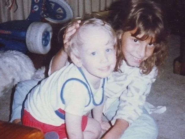 A young boy Coach Brice Williams with curly blonde hair smiling and sitting on a girl with dark brown hair; they are indoors with a skateboard and a dog in the background. Brice A. Williams ; Brice Williams ; Coach Brice Williams.