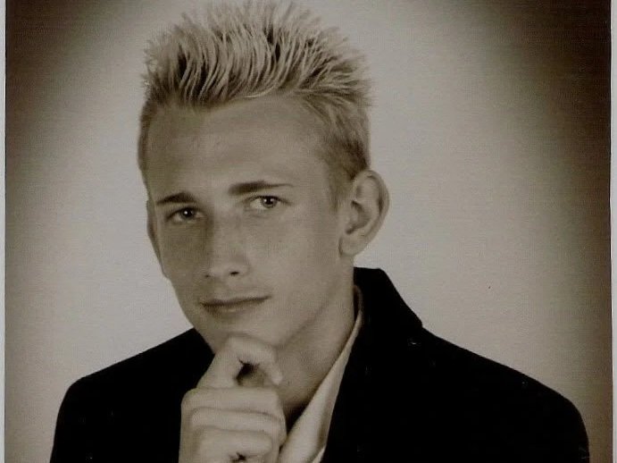 Black and white portrait of a dashing young man with styled frosted tip blonde hair, wearing a dark blazer and resting his chin on his hand, gazing at the camera. Coach Brice Williams ; Brice A. Williams ; Brice Williams. 