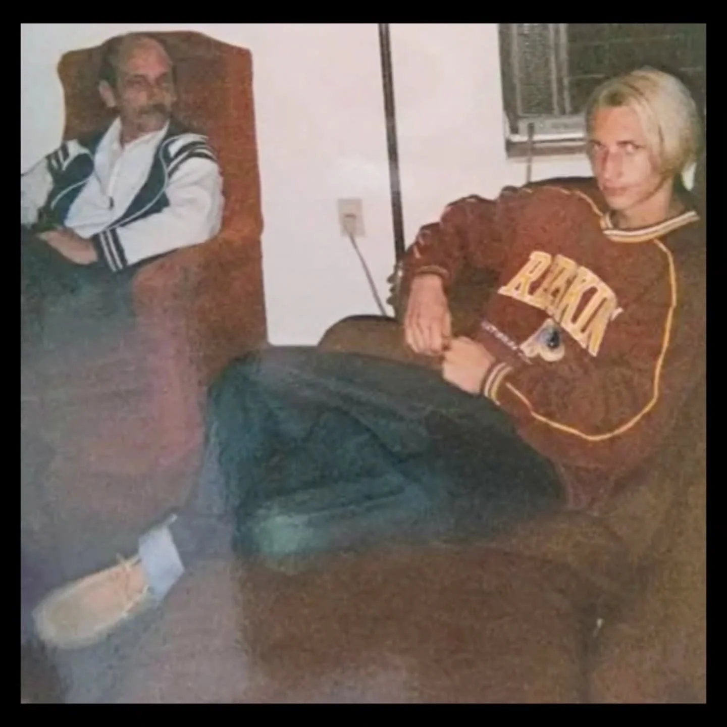 A young man ; Coach Brice Williams, with shoulder-length blonde hair sitting in a living room chair with his Father Richard Williams, one wearing a white jacket with black stripes, the other a red and yellow sports jacket, both looking at the camera.
