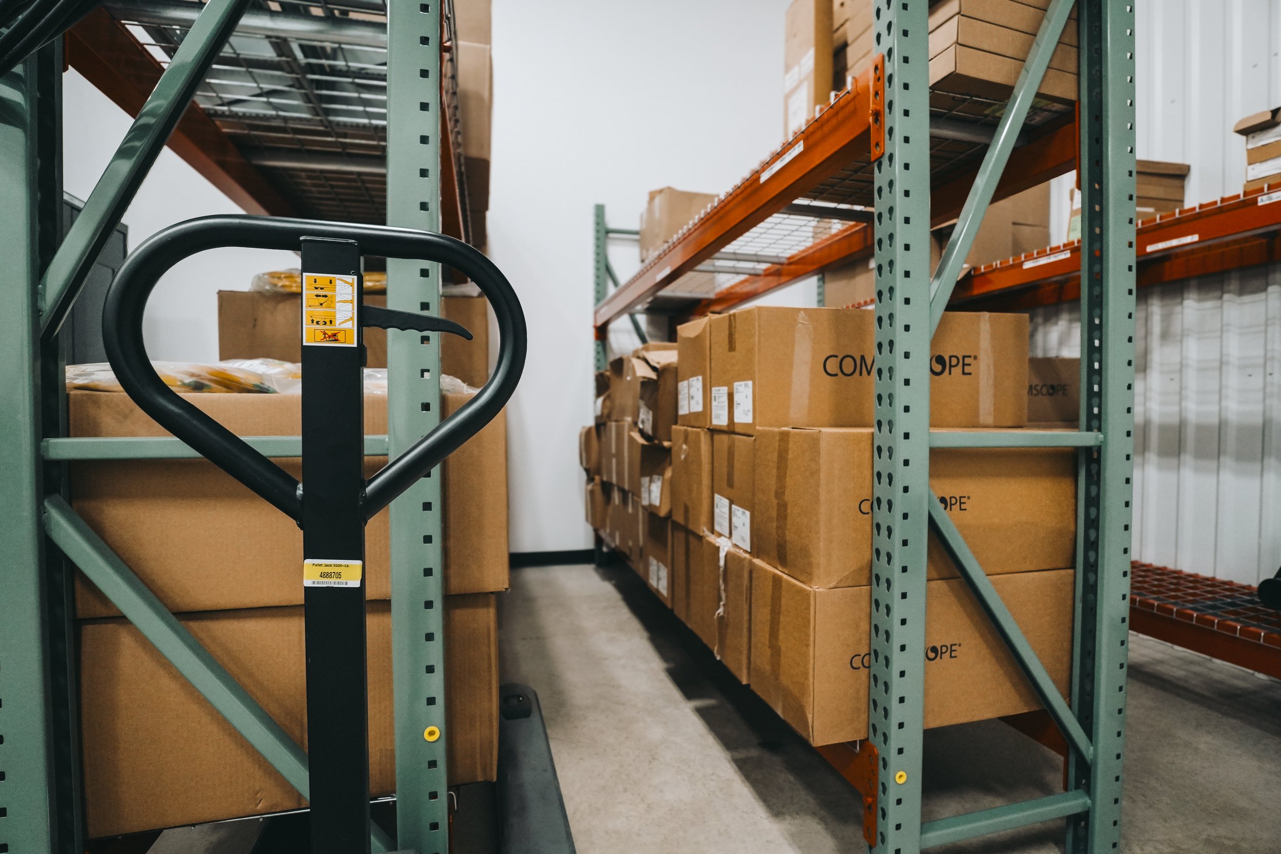 Warehouse shelving filled with packed cardboard boxes, a pallet jack in the foreground, and a white wall in the background.