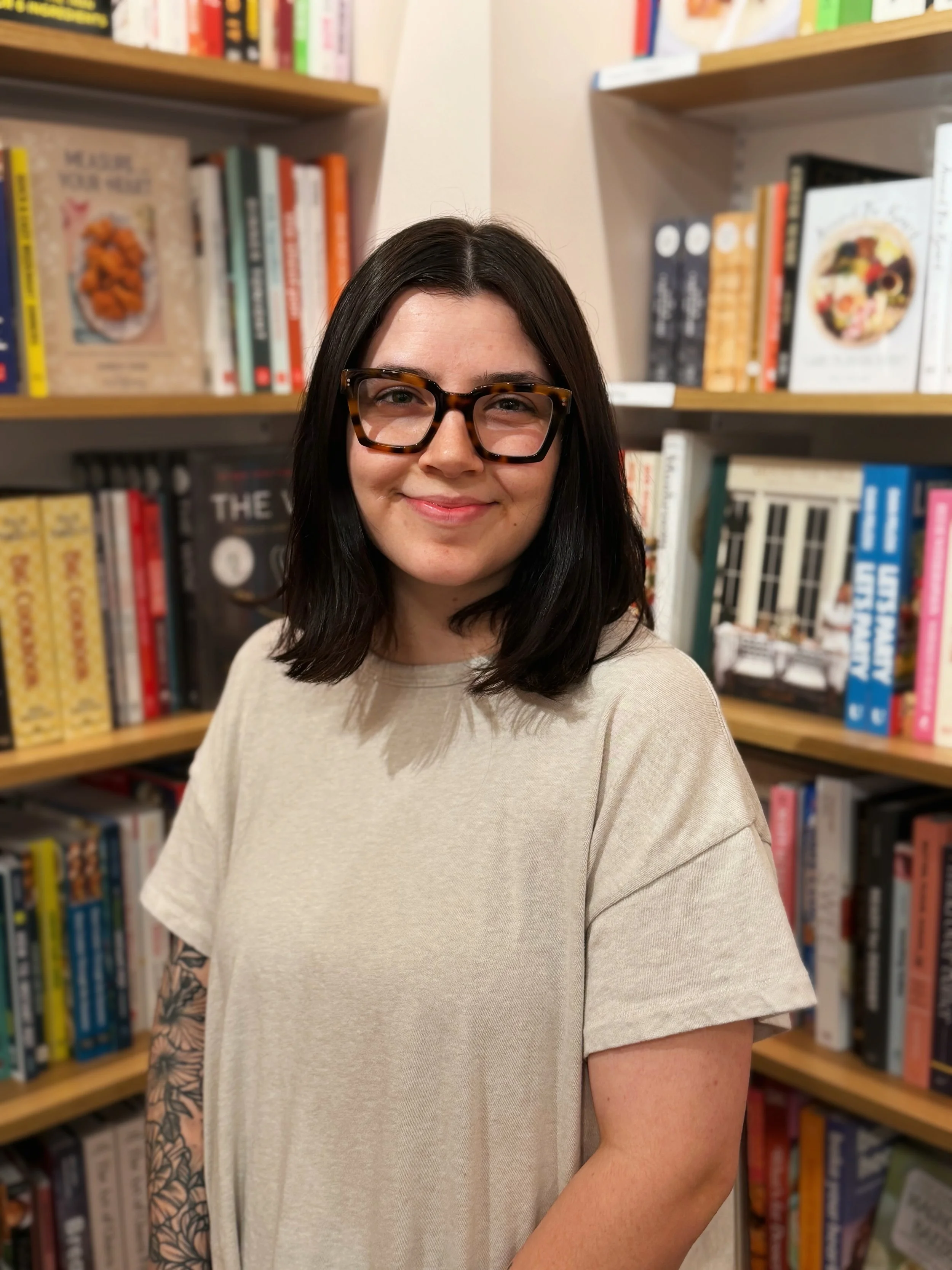 A woman with dark brown hair, glasses, and a light beige t-shirt standing in front of bookshelves filled with colorful books at a library.