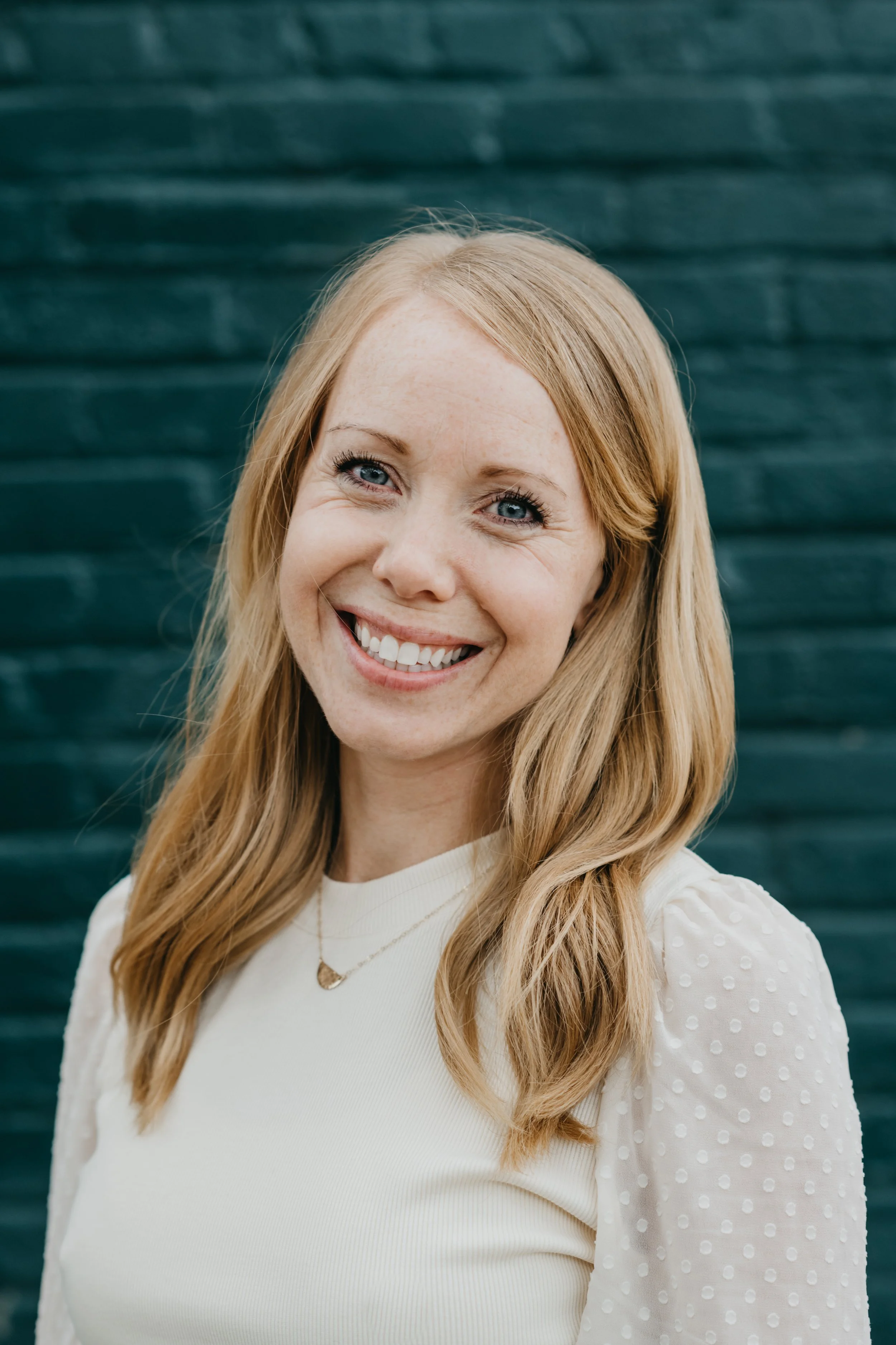 A smiling woman with long red hair wearing a white top with puffed sleeves with dots, standing in front of a dark teal brick wall.