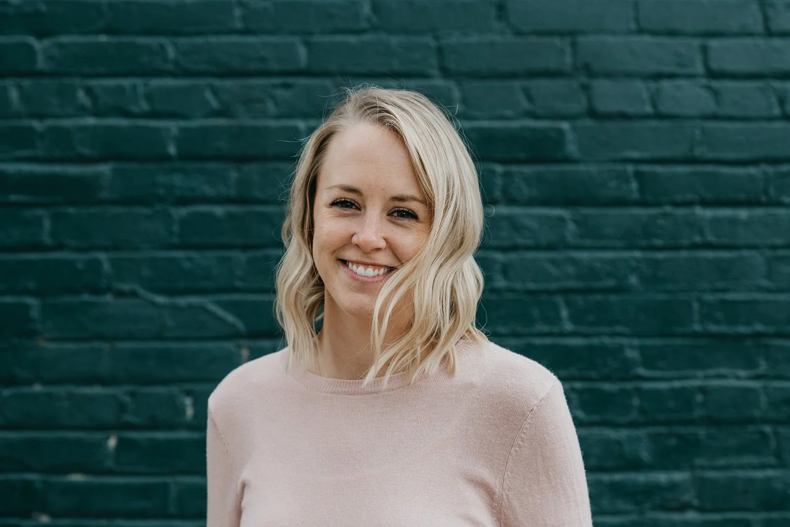 A woman with blonde wavy hair smiling in front of a dark green brick wall.