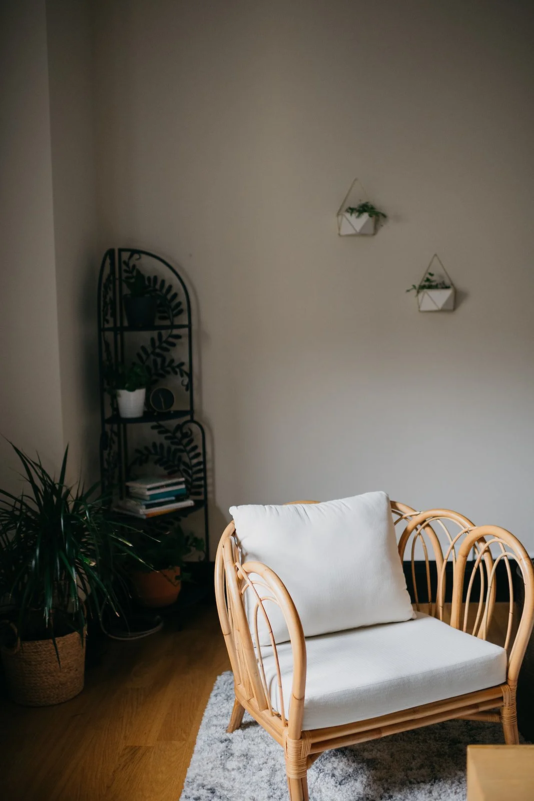 Living room corner with a rattan loveseat, white cushion, potted plants, a black metal shelf with books and decor, and minimalist wall hangings.