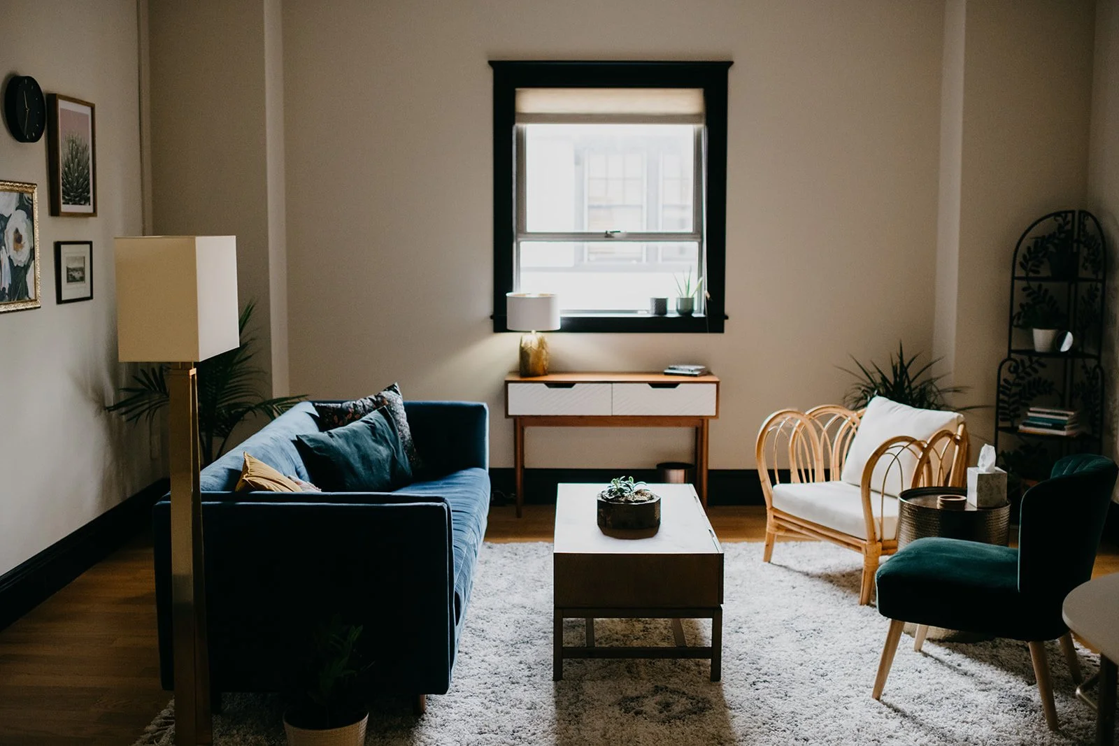 Living room with a blue sofa, a white armchair, a coffee table with a plant, a window, a sideboard with a lamp, and a black metal shelf with plants and books.