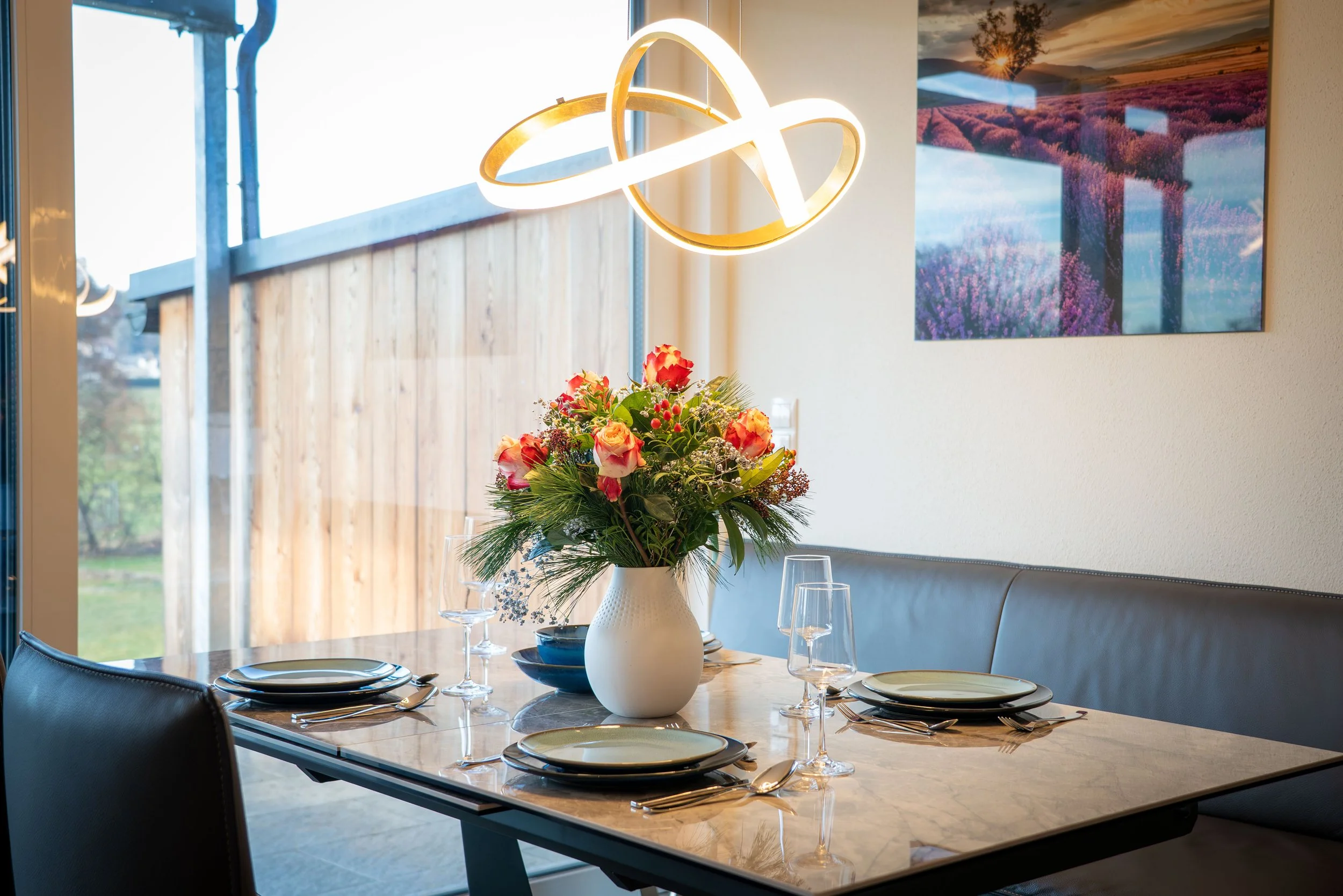 A dining room with a marble table set for four, featuring plates, glasses, and cutlery. A white vase with a bouquet of pink, orange, and yellow flowers sits at the center. In the background, there's a large window with a view of a wooden fence and gr