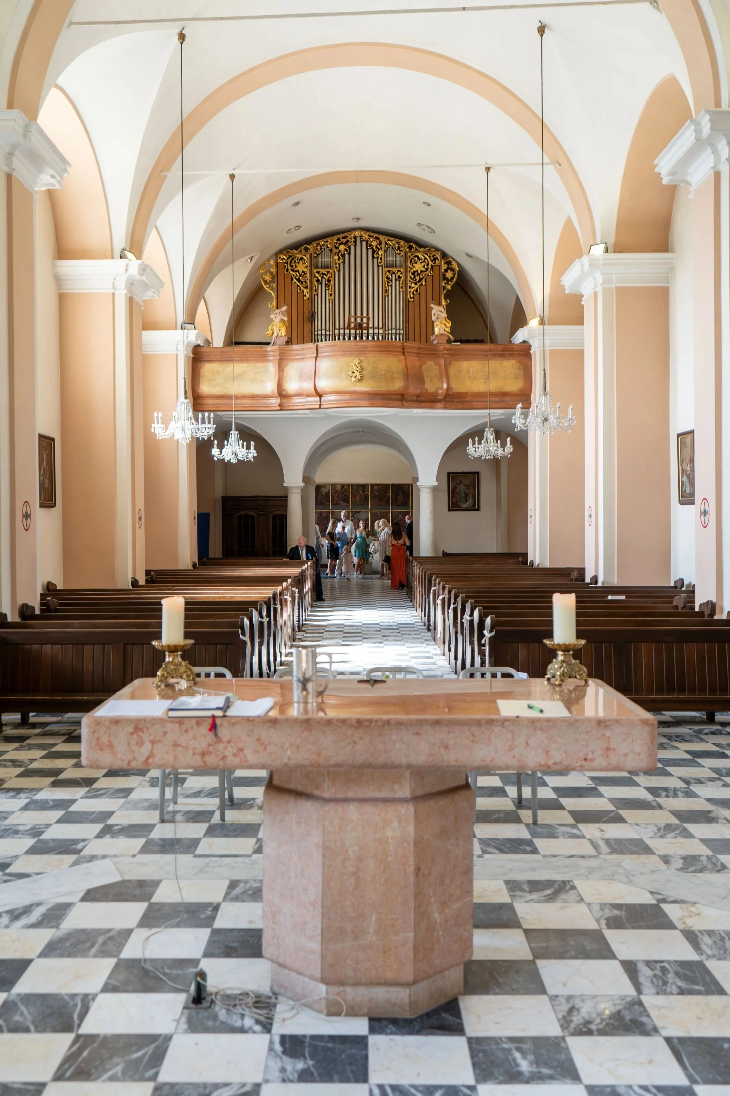 Interior of a church with black and white checkered marble floor, wooden pews, a stone altar with candles, and a high ceiling with arches and chandeliers. In the background, there are several people standing near an altar with religious artwork.