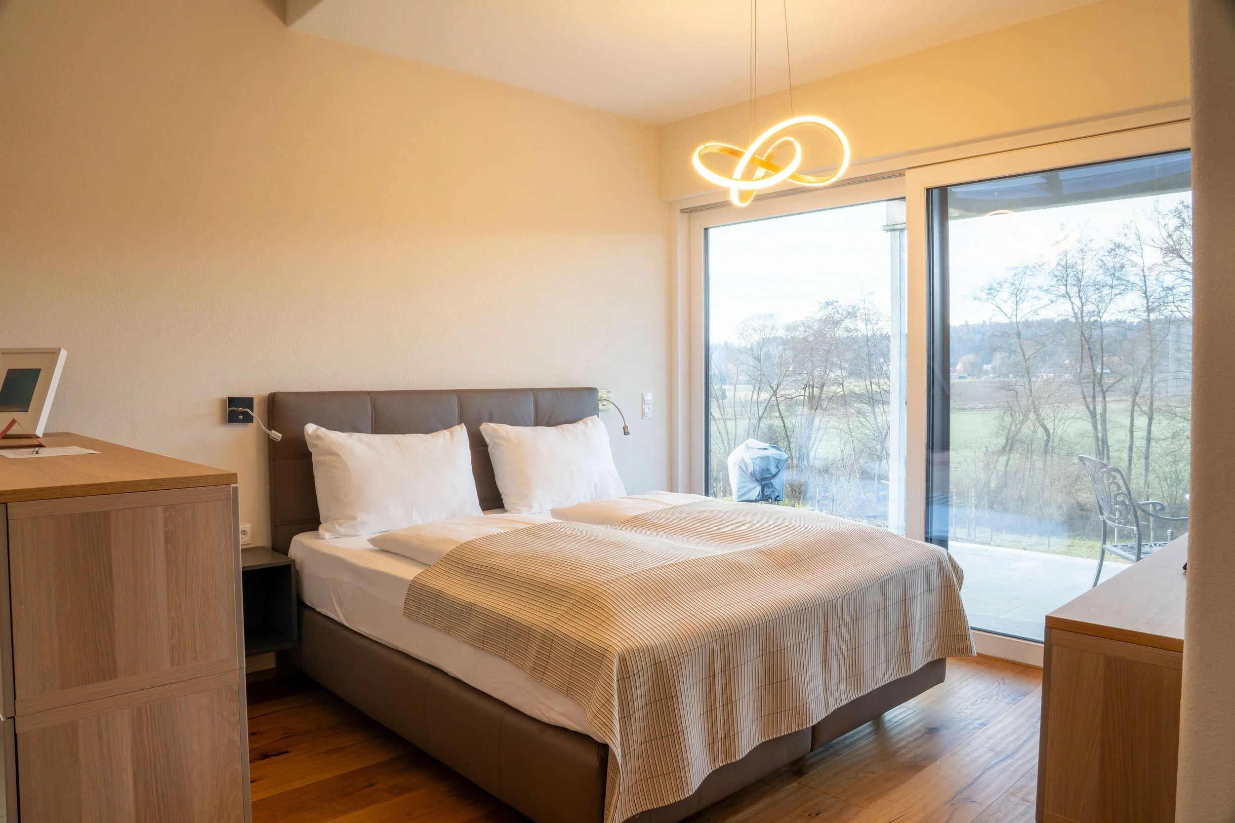 Ein Schlafzimmer mit Doppelbett, weißen Kissen und einer beige-gestreiften Bettdecke, große Fenstertüren mit Blick auf eine natürliche Landschaft im Freien, moderner Kronleuchter an der Decke, Holzfußboden, Tisch mit einem Gerät auf der linken Seite.