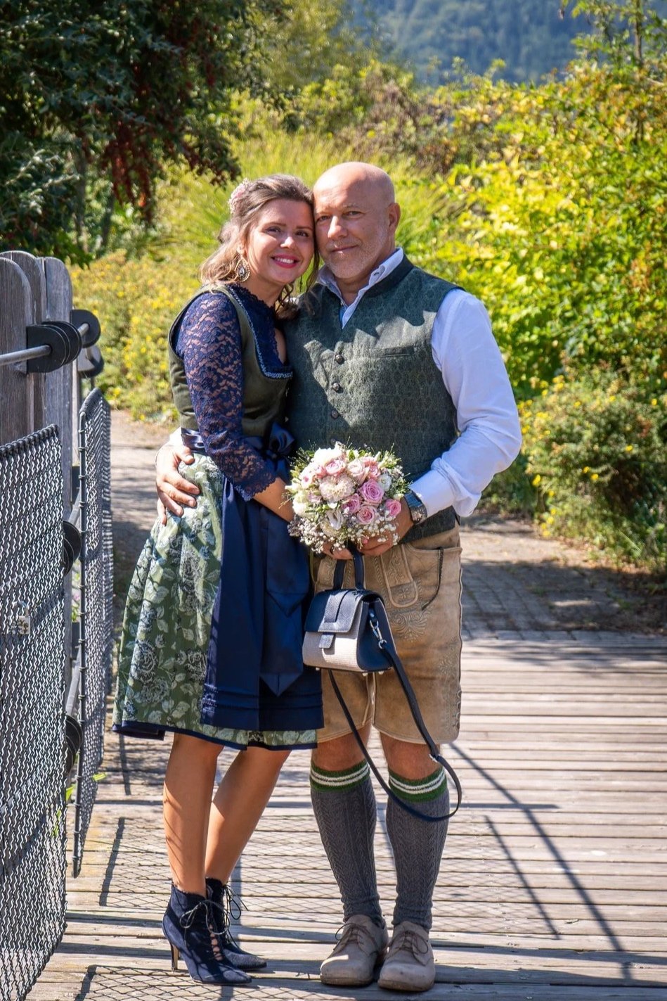 Ein Paar in traditionellen Trachten posiert auf einer Brücke im Freien, die Frau hält einen Blumenstrauß, im Hintergrund bunte Blätter und Bäume.
