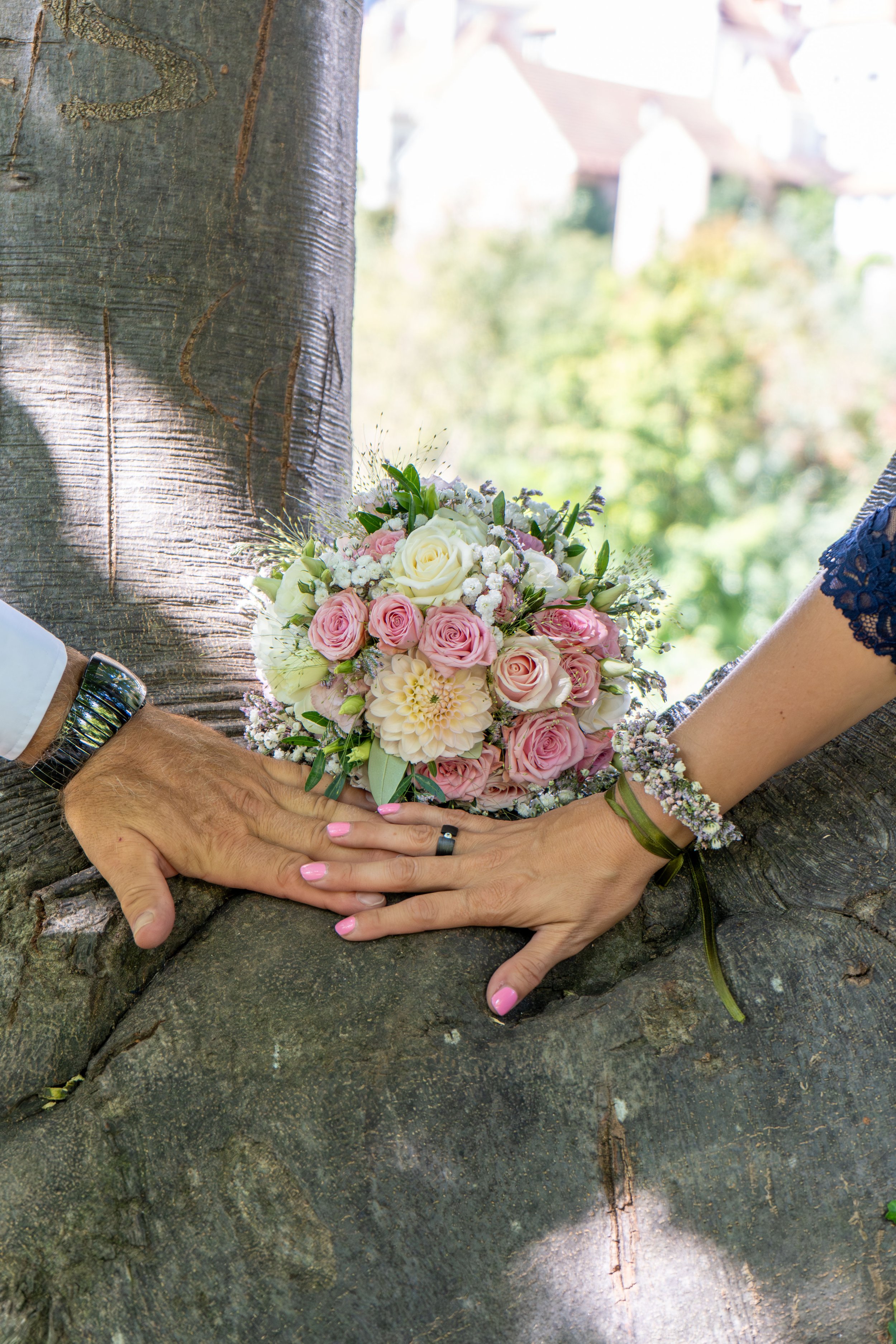 Nahaufnahme eines Brautpaares, das ihre Hände hält und einen Blumenstrauß zwischen ihren Händen hält. Das Bild zeigt nur die Hände, Arme und die Blumen. Der Hintergrund ist verschwommen mit einer grünen Baumlandschaft.