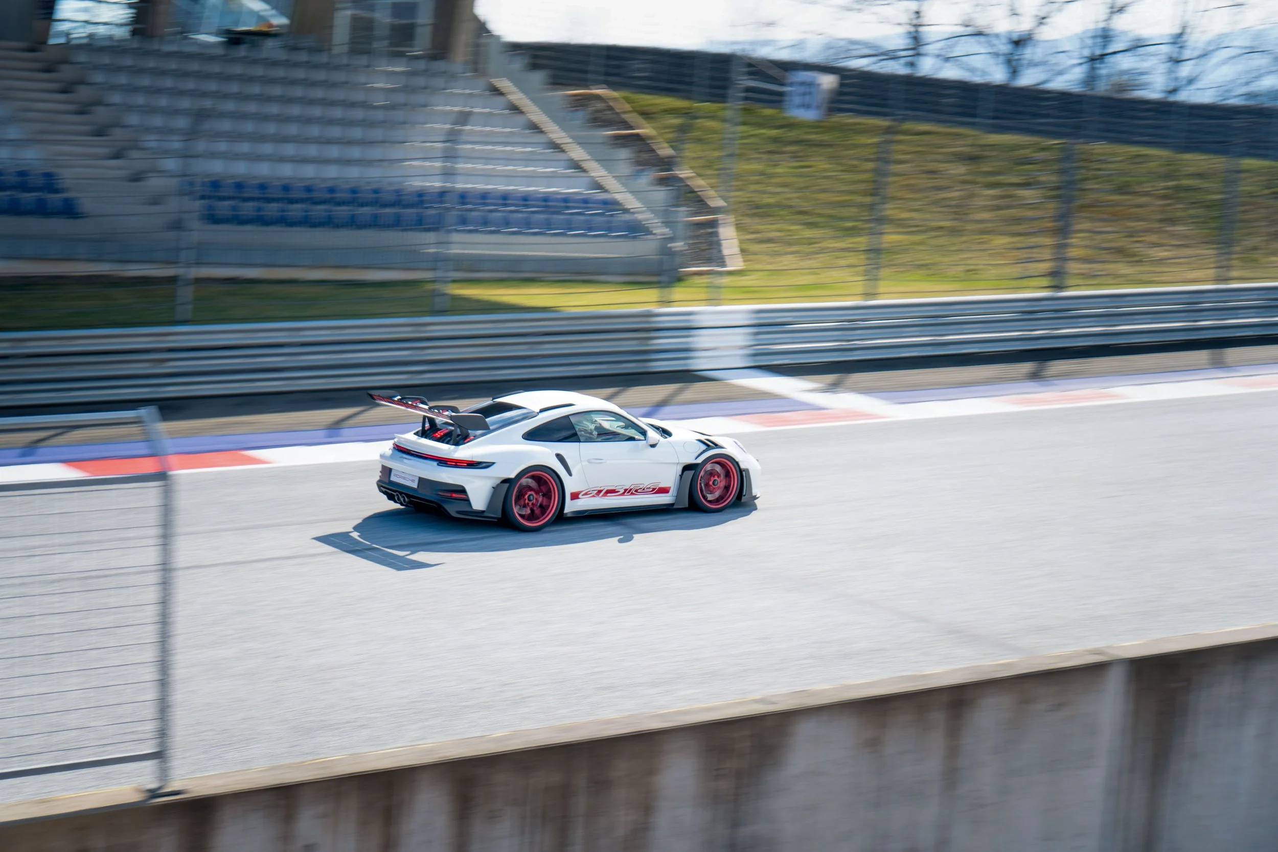 A white sports car with red accents driving on a racetrack, blurred background of empty grandstands and fencing.