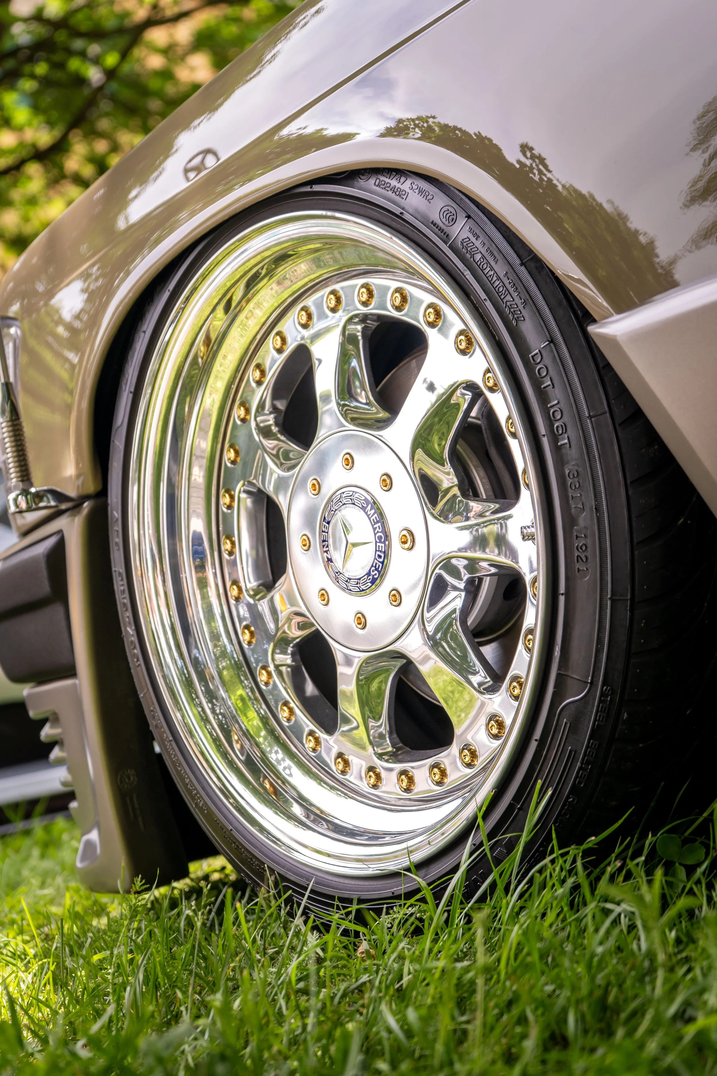 Close-up of a Mercedes-Benz car wheel with a chrome rim and gold accents, parked on grass with trees reflected on the car body.