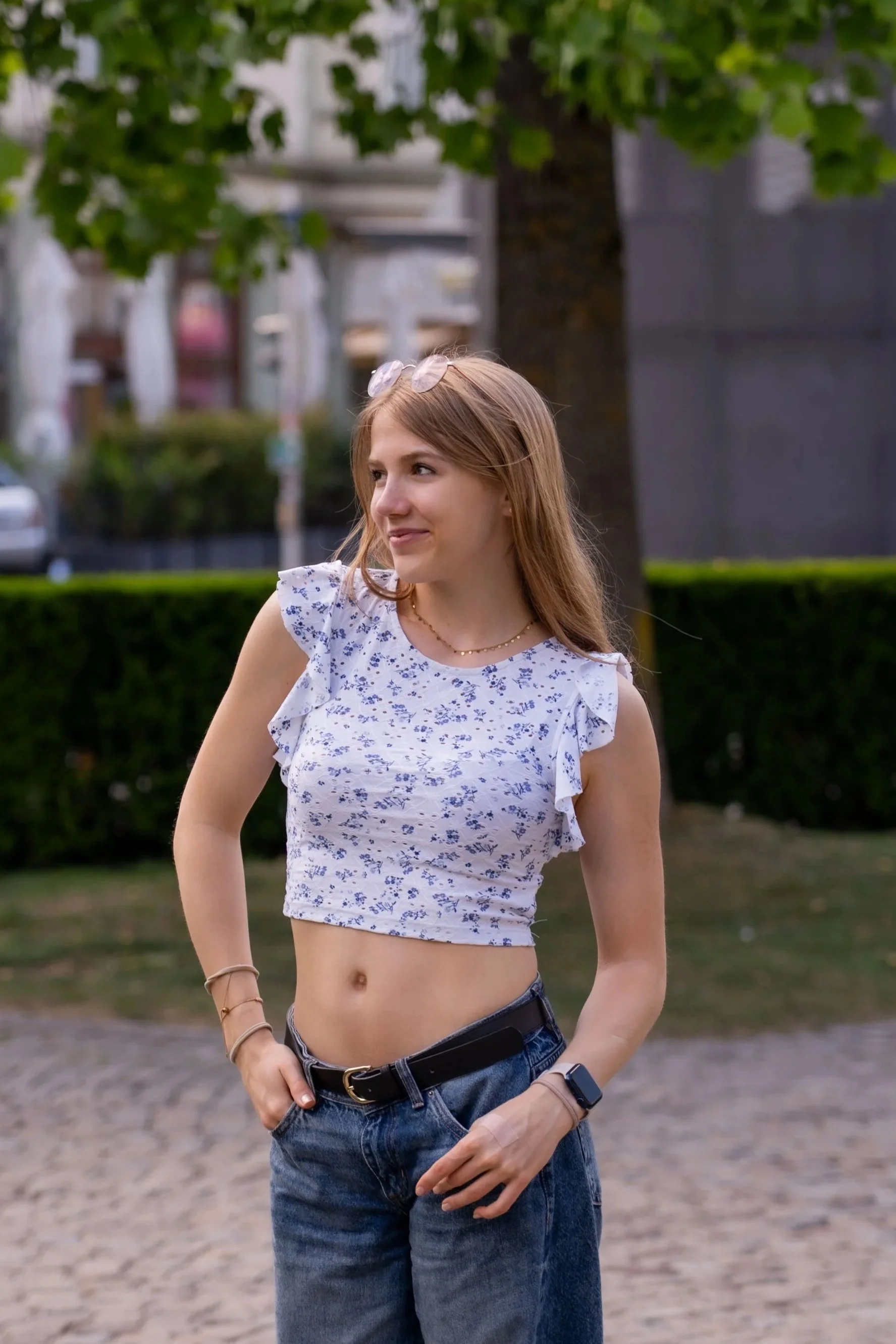 Junge Frau mit roten Haaren, Sonnenbrille auf dem Kopf, trägt ein weißes Top mit blauen Blumenmuster, Jeans, in einer parkähnlichen Umgebung mit Bäumen und Zäunen.