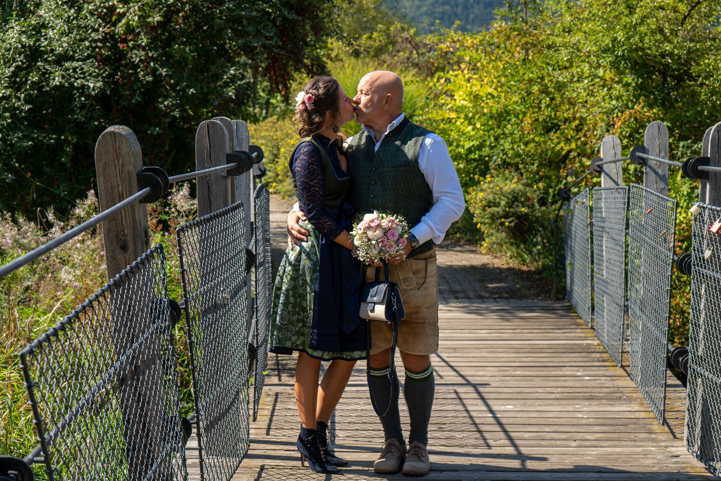 A couple sharing a kiss on a wooden bridge surrounded by lush greenery, woman holding a bouquet of pink roses.
