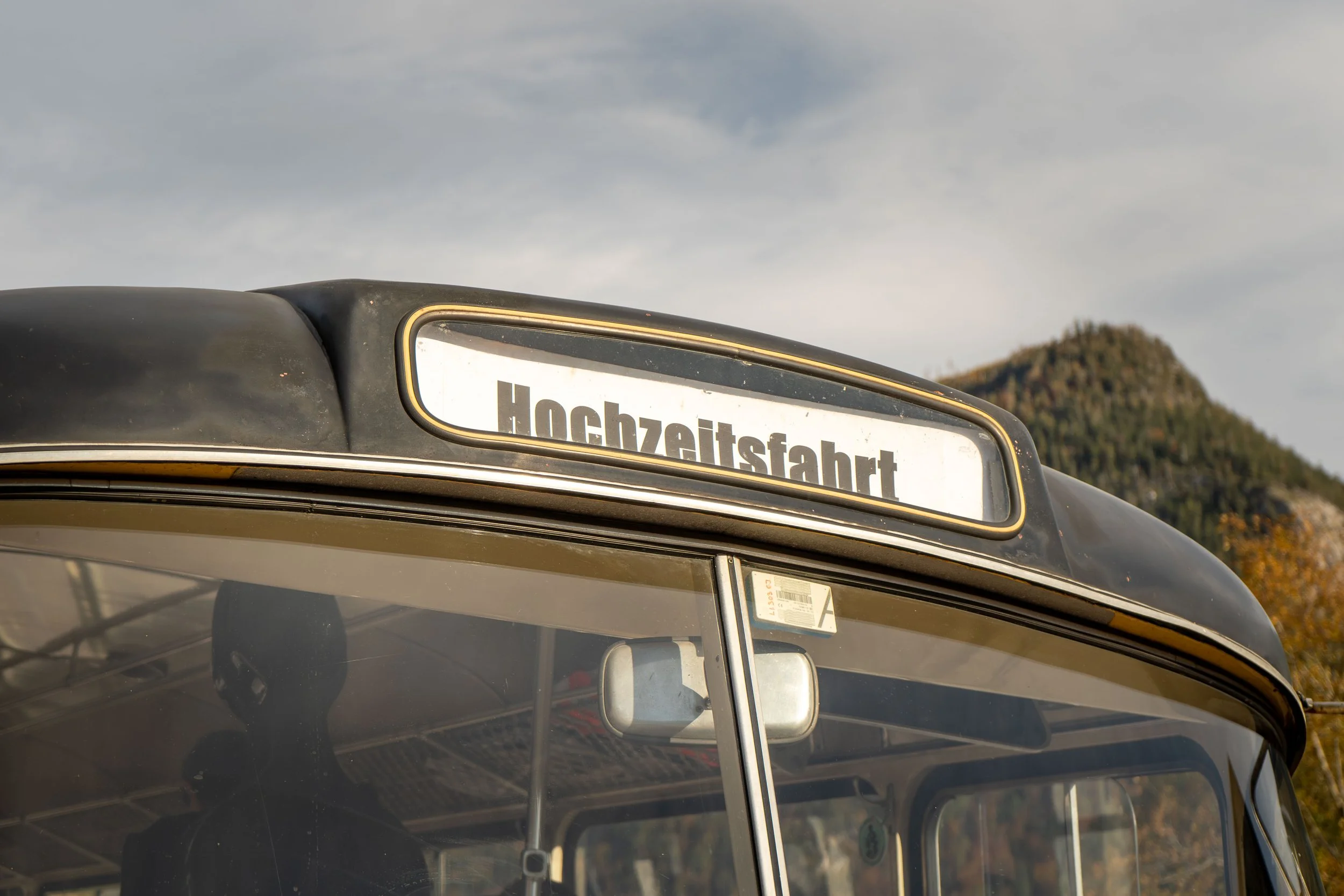 Tourist bus with destination sign reading "Hochzeitsfahrt" parked outdoors, with a mountain in the background and cloudy sky.