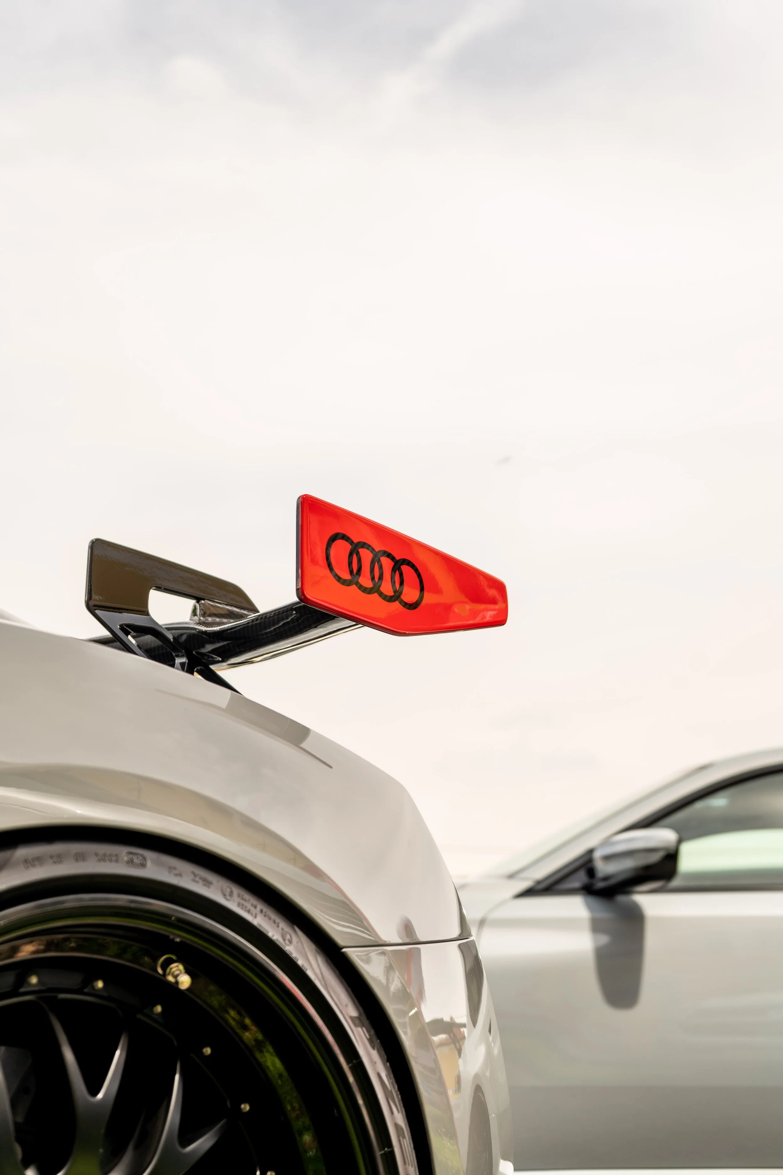 Vorderansicht von mehreren Autos, Fokus auf einen weißen Audi mit rotem Heckspoiler, über dem ein grauer Himmel.
