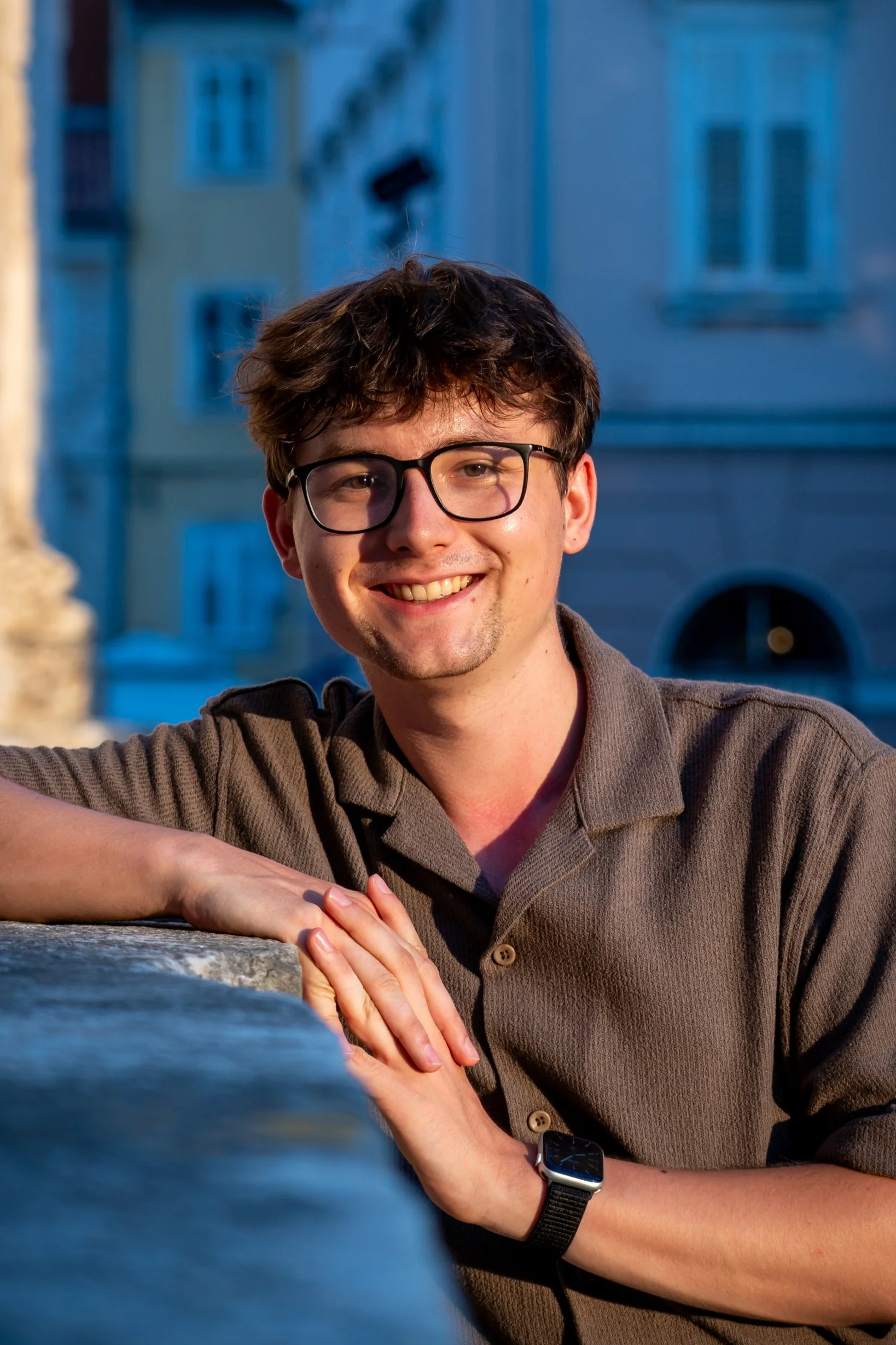 Junge Mann mit Brille und Smartwatch, lächelnd, am Abend, im Freien, mit historischen Gebäuden im Hintergrund.