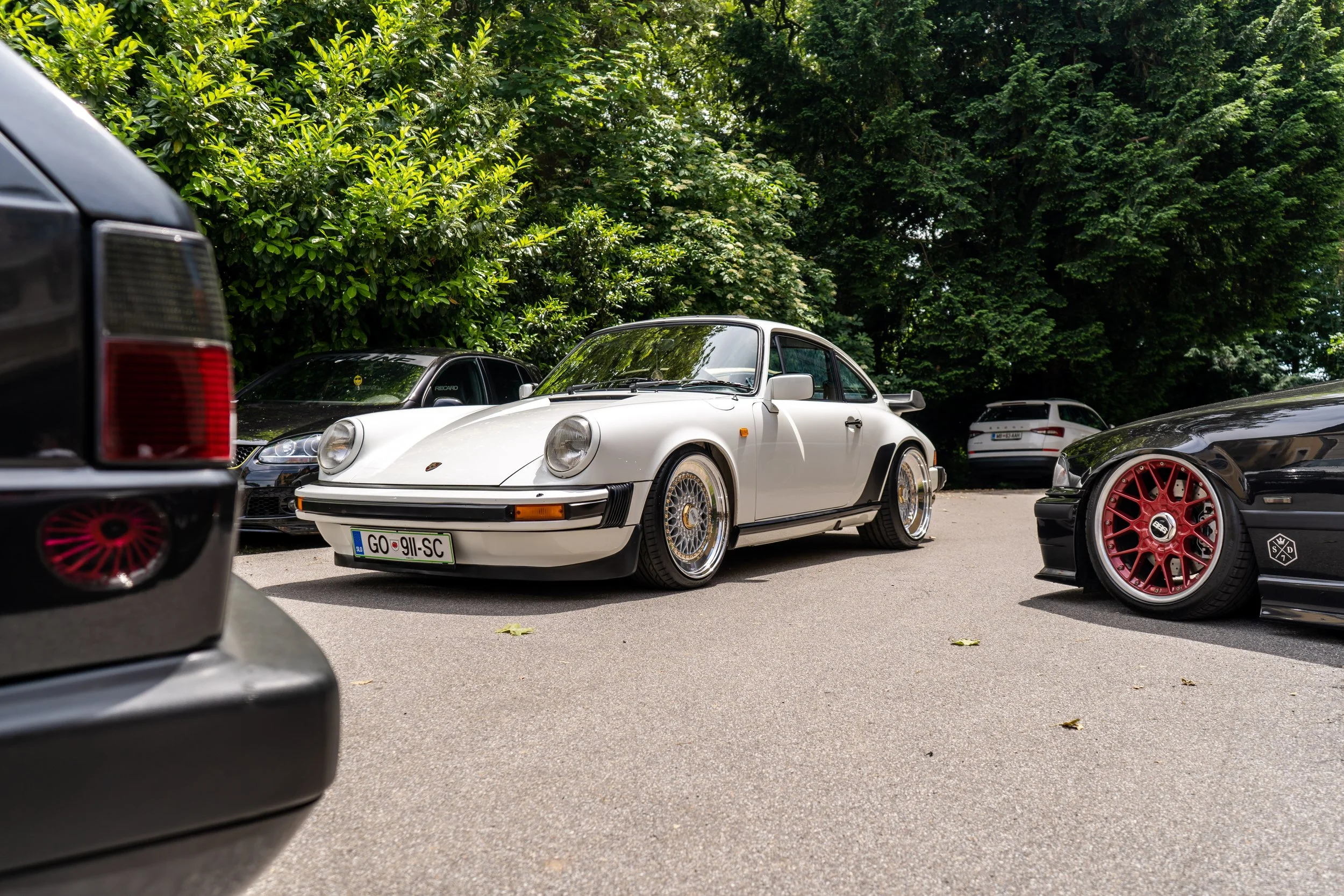 Ein weißer Porsche 911 mit individuellen Felgen steht auf einem Parkplatz, umgeben von anderen Autos und dichter grüner Baumvegetation im Hintergrund.